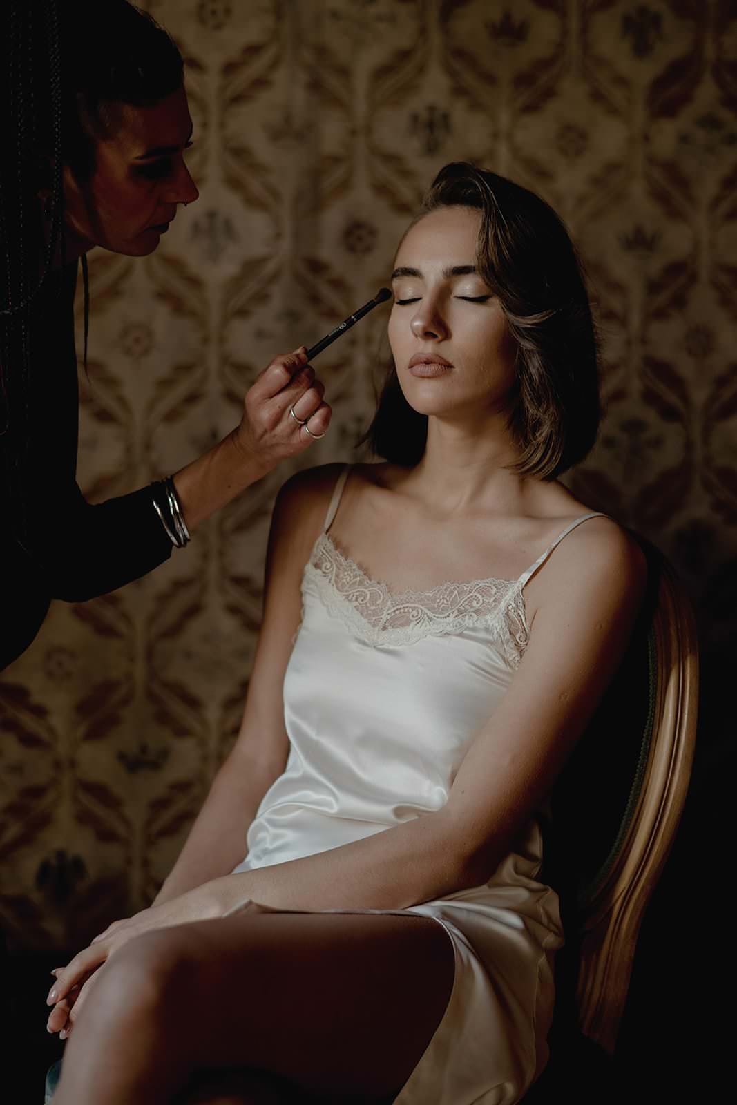 A woman in a Grace Loves Lace Suki dress sits with her eyes closed as a person applies makeup to her eyelids. The scene, reminiscent of a vintage wedding in Tuscany, features patterned wallpaper and soft, ambient lighting. Tuscan Wedding Photographer