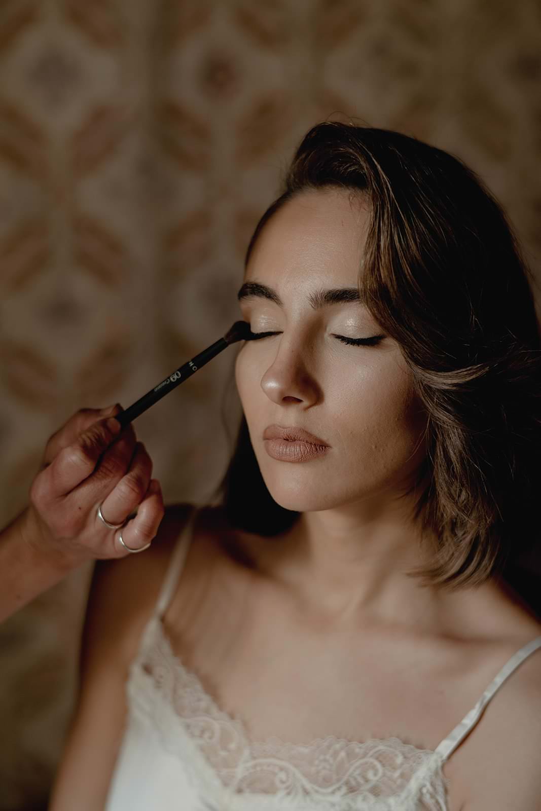 A woman in a white lace-trimmed top, reminiscent of a Grace Loves Lace design, has her eyes closed as a hand expertly applies eye makeup. The soft, blurred background evokes the calm ambiance of a wedding in Tuscany. Her hair falls in loose waves, and her expression is serene. Tuscan Wedding Photographer