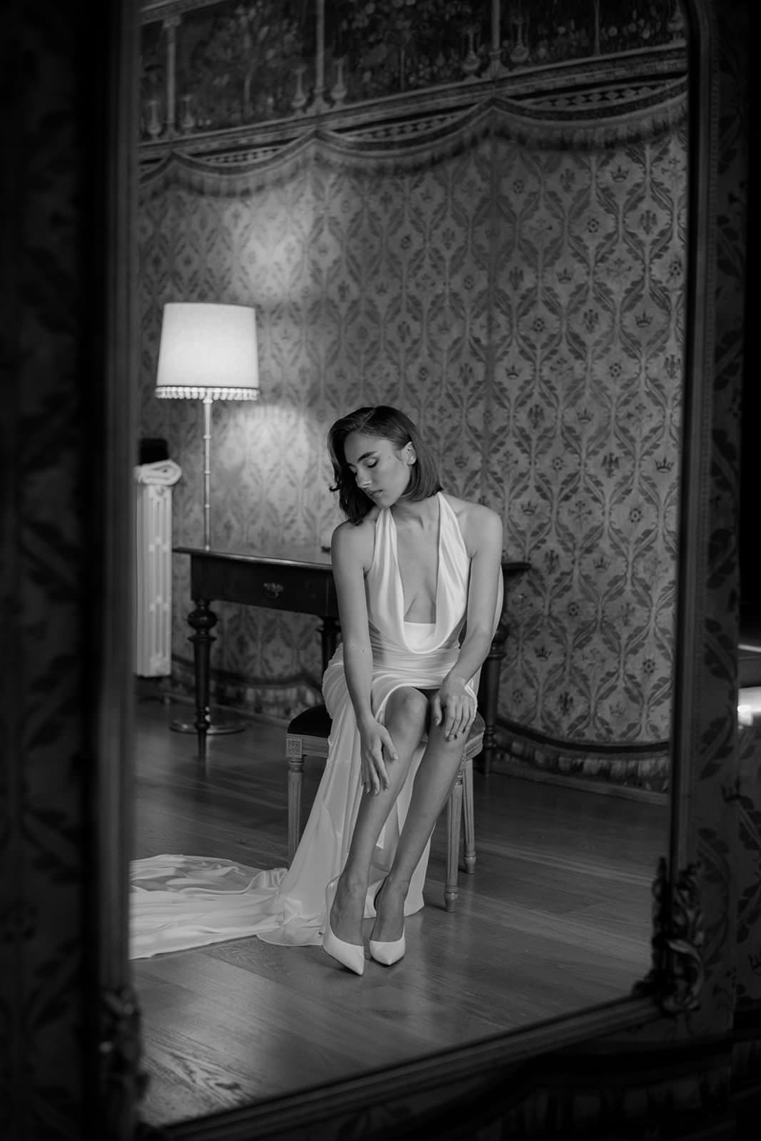 A woman in a flowing Grace Loves Lace Suki dress sits on a chair in an elegantly decorated room. She gazes down thoughtfully, with her hands resting on her knees. The setting features patterned wallpaper, a standing lamp, and a framed mirror capturing her reflection. The image is in black and white. Tuscan Wedding Photographer
