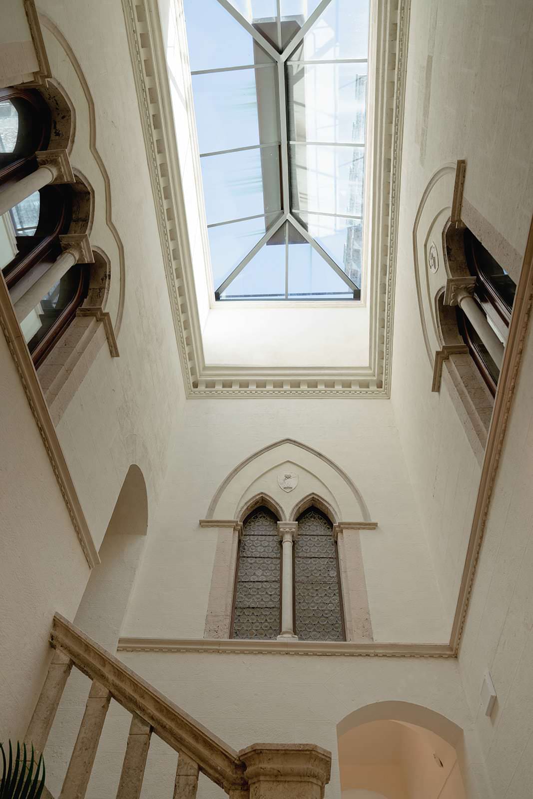 View of a high-ceiling interior at Palazzo al Piano, a stunning wedding location in Tuscany. The skylights geometric design allows natural light to flood the room, highlighting arched windows and white walls. Ornate stone details and an elegant staircase add historical charm to this bright, airy setting. Tuscan Wedding Photographer