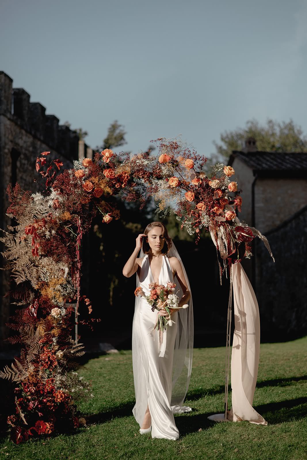 The bride, in a stunning Grace Loves Lace Suki dress, stands under an arch adorned with vibrant flowers and greenery. She holds a bouquet on a grassy landscape with stone buildings in the background. The warm, natural light enhances the romantic atmosphere of this picturesque wedding in Tuscany. Tuscan Wedding Photographer