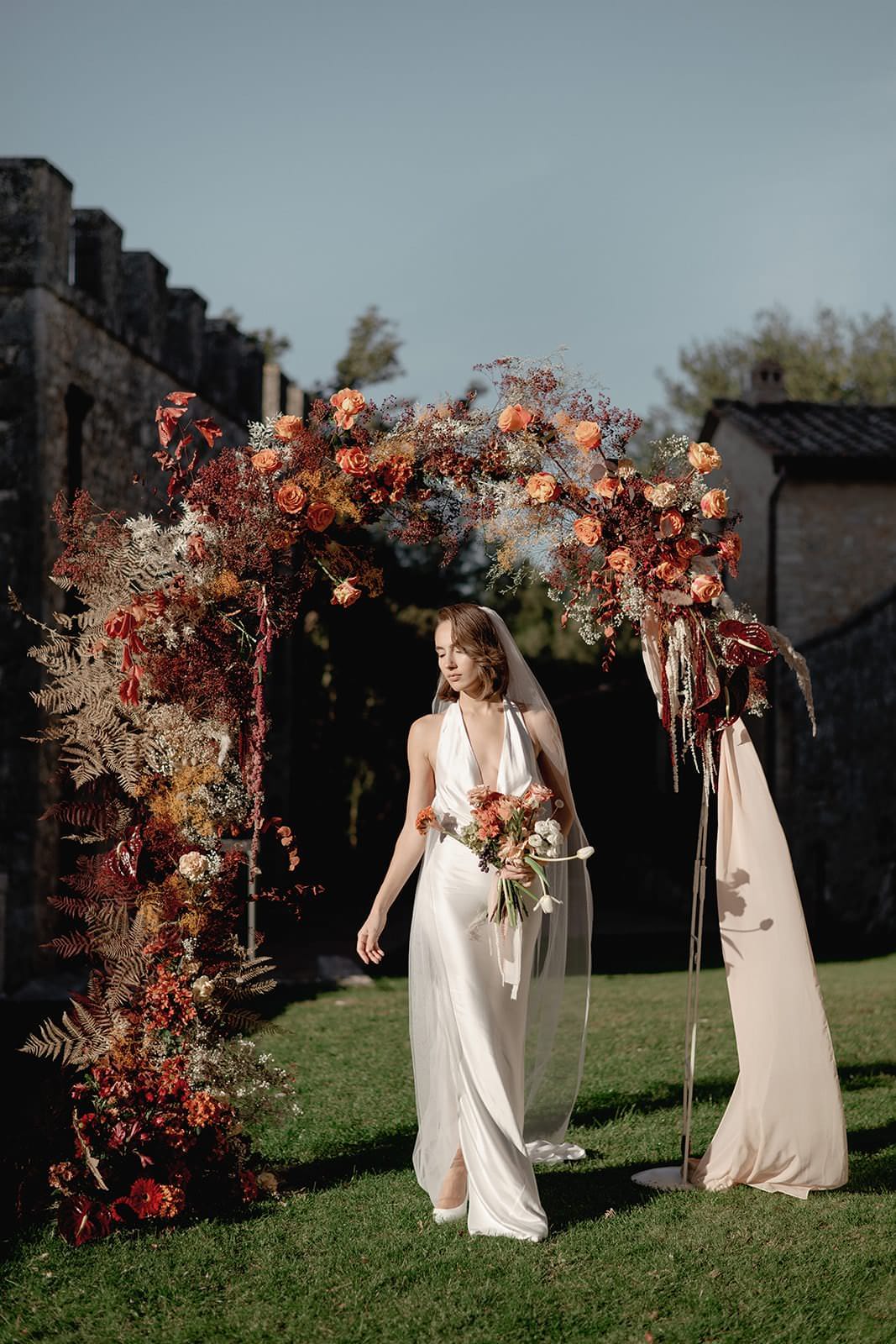 A bride, shimmering in a sleek Grace Loves Lace Suki Dress, holds a bouquet under a rustic floral arch. Vibrant red, orange, and yellow blooms with greenery frame her against an old stone structure. This outdoor wedding in Tuscany unfolds beneath a clear and bright sky. Tuscan Wedding Photographer