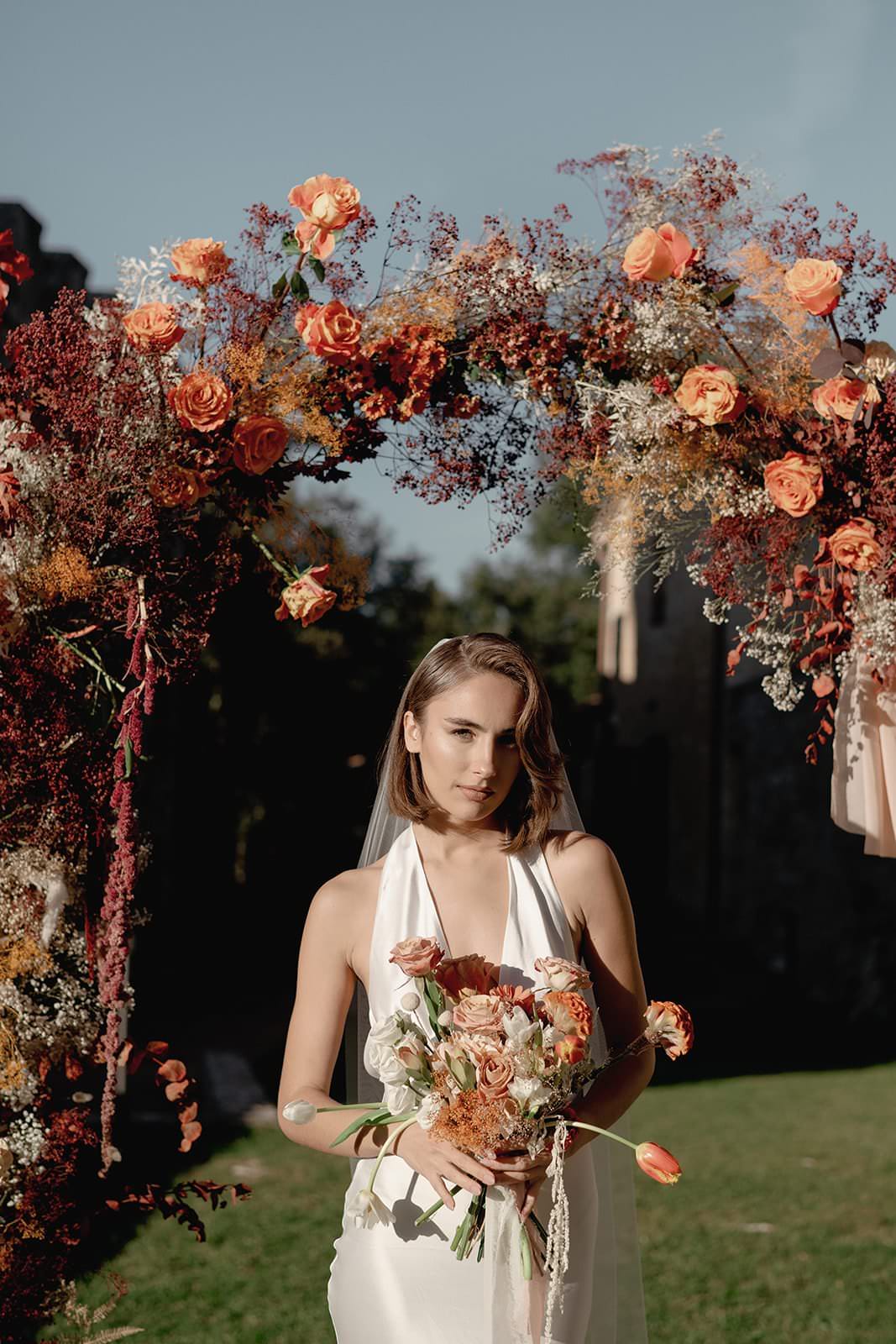 A woman in a Grace Loves Lace Suki Dress stands under a floral arch adorned with orange and red flowers. Holding a matching bouquet, she is set against the sunny outdoor backdrop of a Tuscany wedding, complete with green grass and trees that create a warm, romantic atmosphere. Tuscan Wedding Photographer