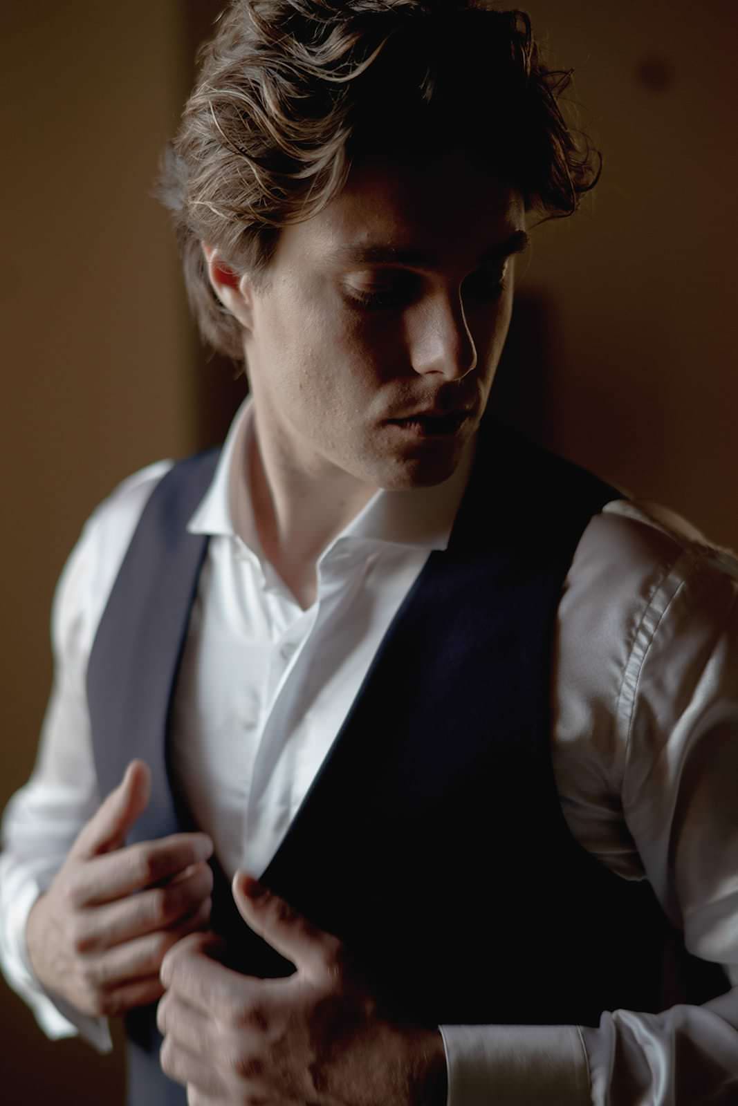 A man with wavy brown hair is adjusting his dark vest over a white dress shirt. He is looking downward to his left, standing in a softly lit room with a shadowed background at Palazzo al Piano, an elegant wedding location in Tuscany. The image captures a moment of quiet concentration. Tuscan Wedding Photographer