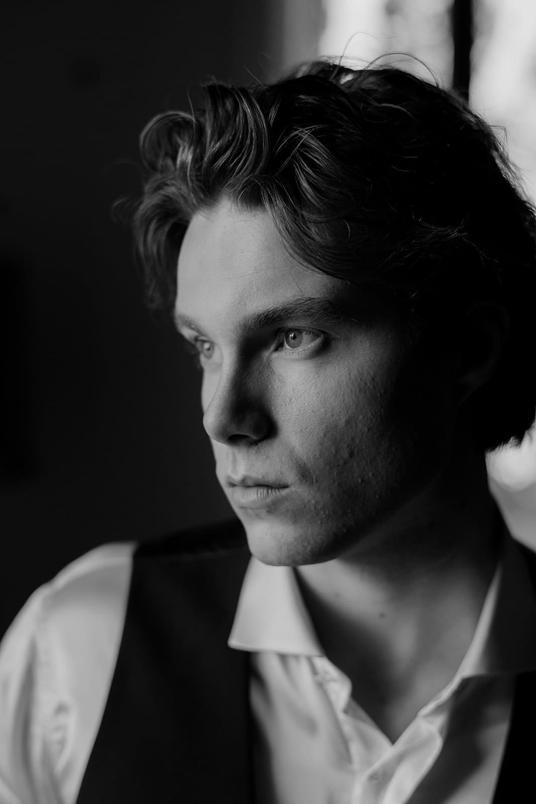 Black and white portrait of a young man with wavy hair, looking thoughtfully to the side. He wears a white shirt and dark vest; soft, natural light highlights his face, casting gentle shadows. The moody atmosphere evokes the charm of a Palazzo al Piano wedding location in Tuscany. Tuscan Wedding Photographer
