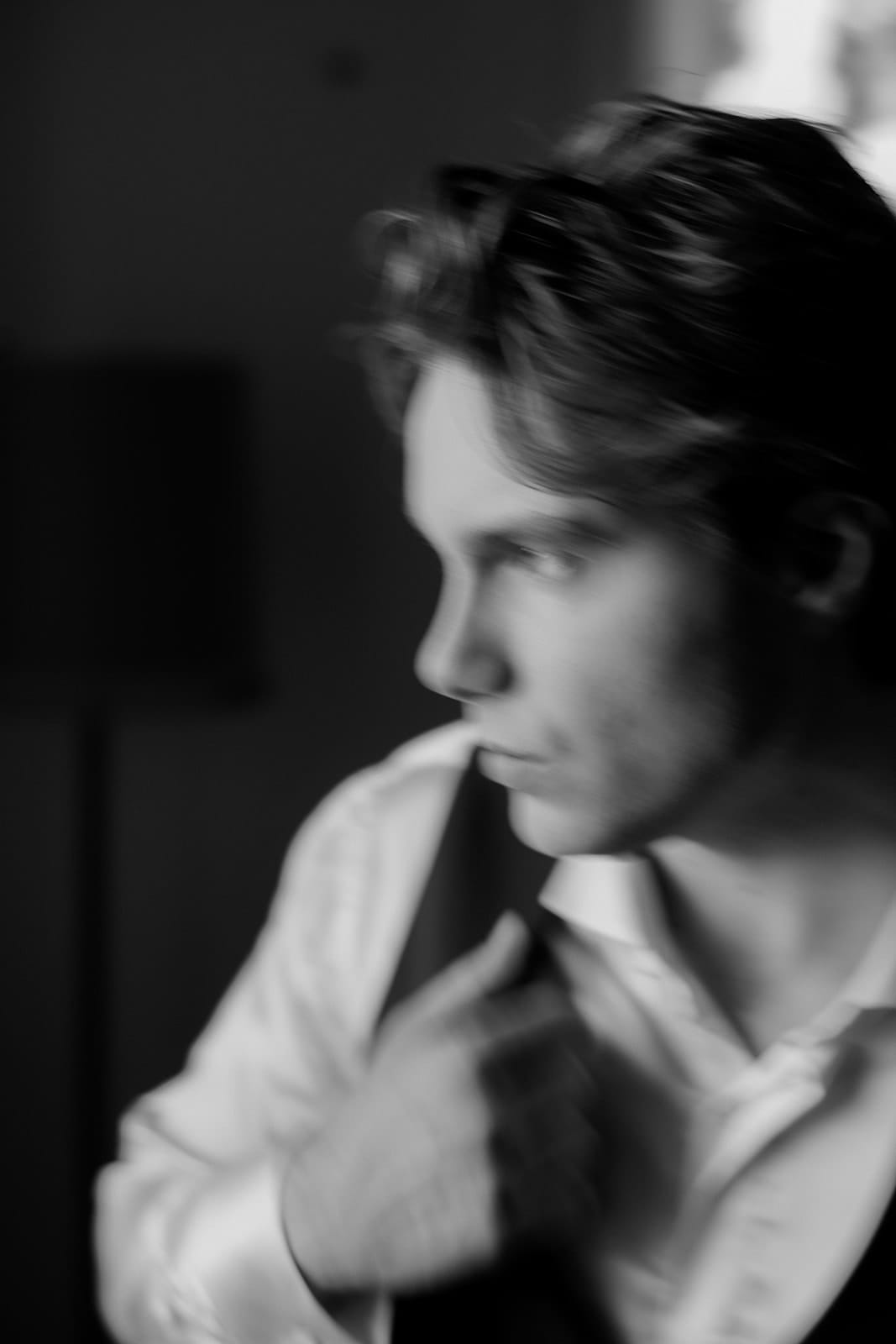 In a black and white photo, a person wearing a white Grace Loves Lace shirt, with medium-length hair, is captured in side profile. They adjust their collar with a graceful touch of the right hand. The background, softly blurred, suggests the elegance of Tuscany. Tuscan Wedding Photographer