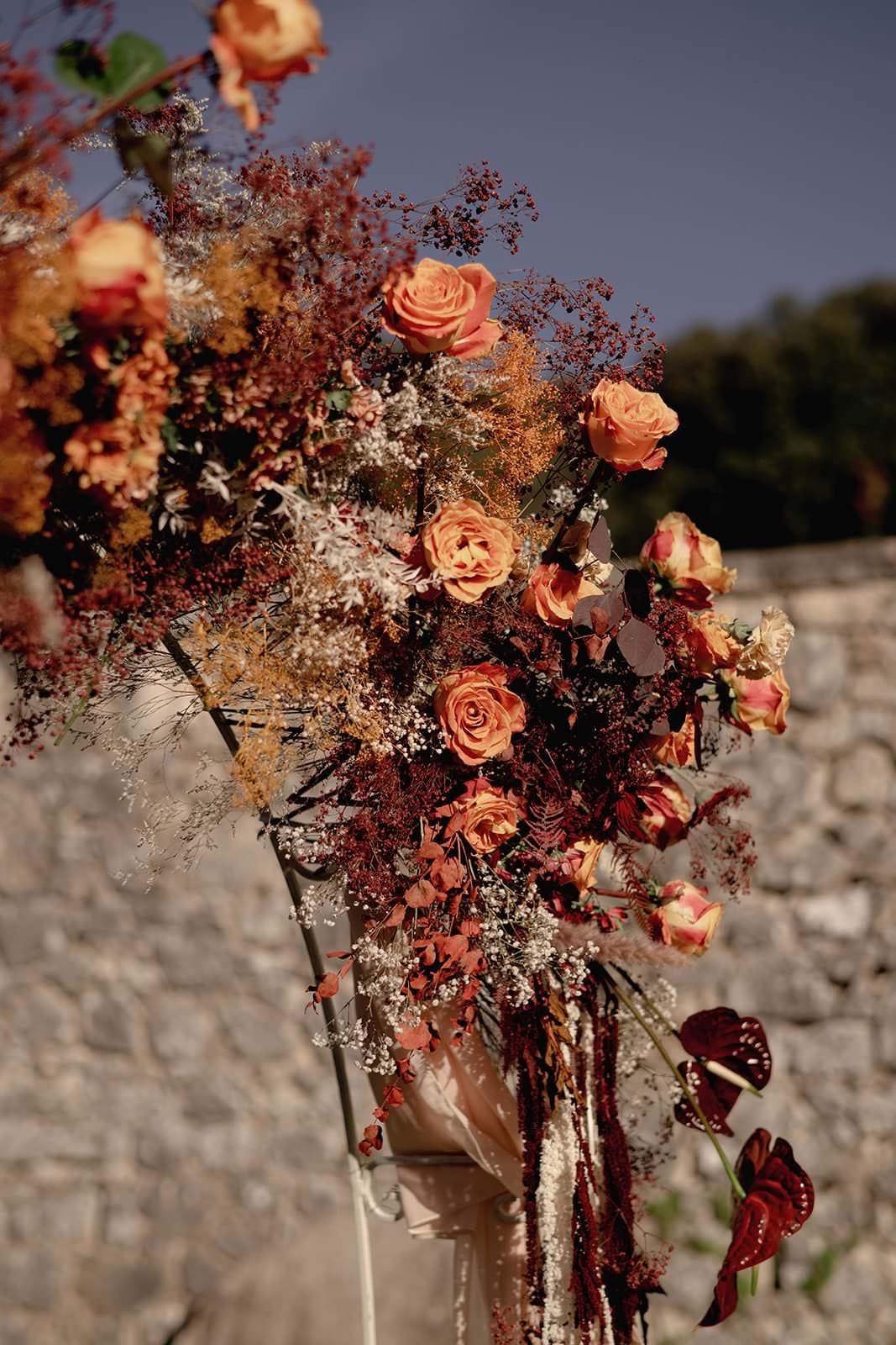 Elegant floral arrangement featuring orange and peach roses, surrounded by dried foliage and greenery, beautifully complementing a Grace Loves Lace Suki dress. Set against a blurred stone wall and trees under a clear blue sky, the scene captures an autumnal wedding in Tuscany ambiance. Tuscan Wedding Photographer
