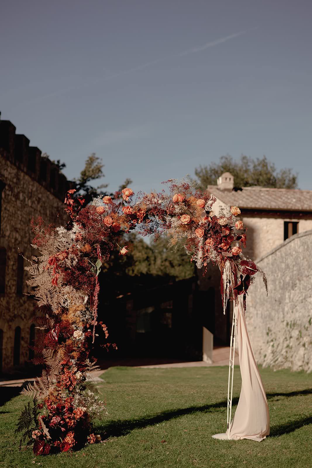 A wedding arch outdoors adorned with vibrant autumn flowers in shades of red, orange, and pink complements the elegance of a Grace Loves Lace Suki dress. Set on a grassy area near a rustic stone building, the archs draped beige fabric enhances this sunny day in Tuscany. Tuscan Wedding Photographer