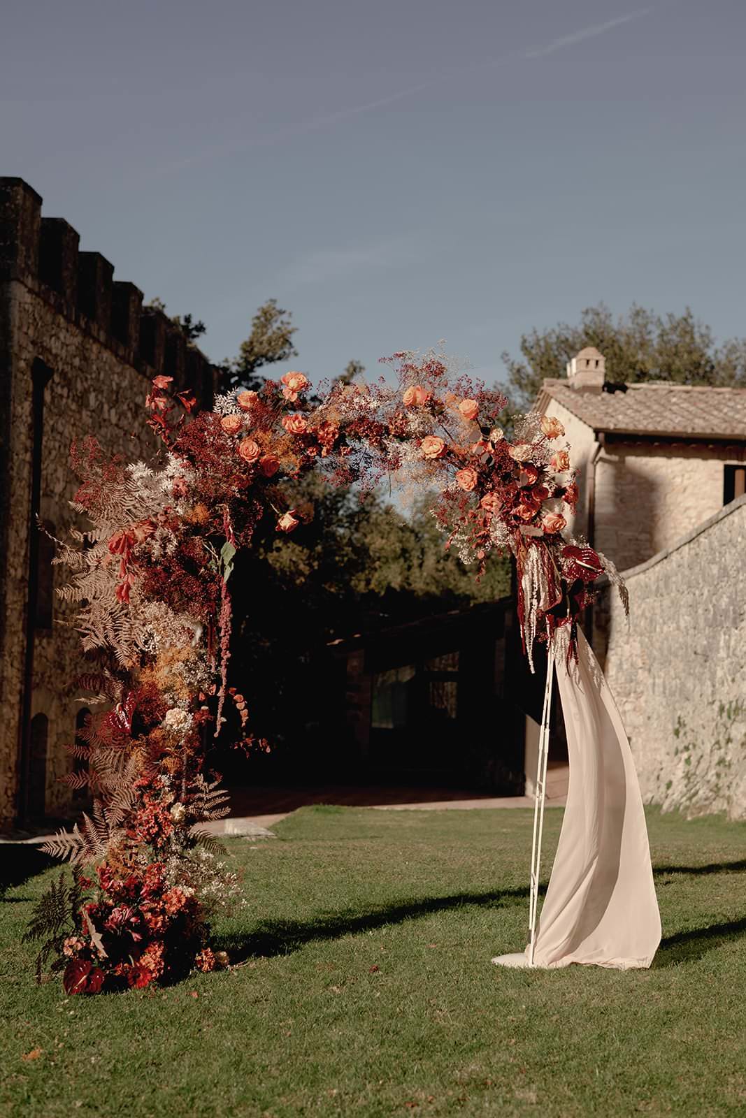 An outdoor wedding arch adorned with deep red and orange flowers, greenery, and flowing fabric stands on lush green grass. Against the castle-like stone buildings of a Tuscany wedding, beneath a clear blue sky, sunlight casts soft shadows for a warm, romantic atmosphere. Tuscan Wedding Photographer