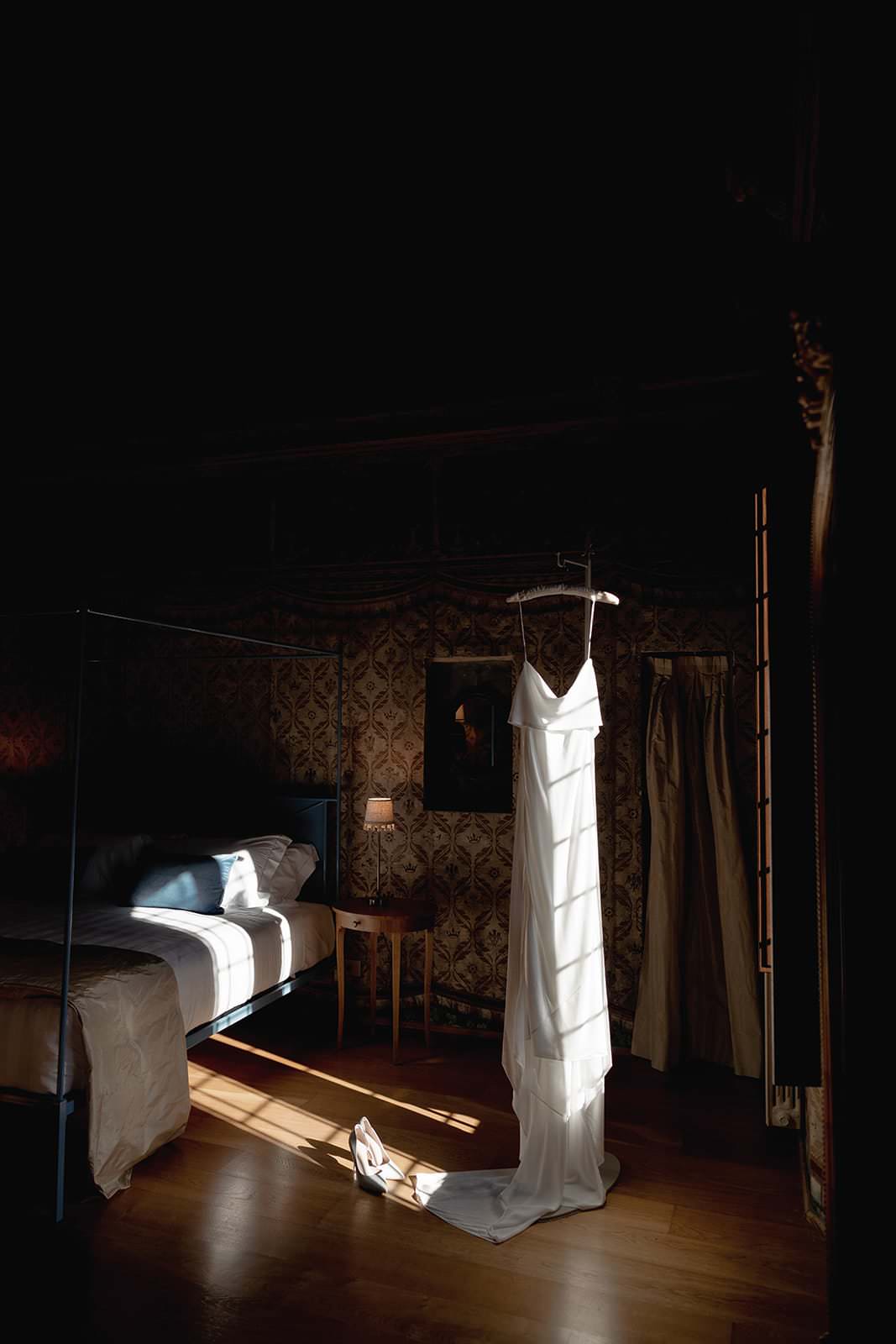 A white wedding dress hangs elegantly from a hanger in a dimly lit bedroom at the Palazzo al Piano wedding location in Tuscany, with sunlight streaming through a window. The dress casts a shadow on the wooden floor, beside a pair of white heels. A bed and table with a lamp adorn the softly lit room. Tuscan Wedding Photographer