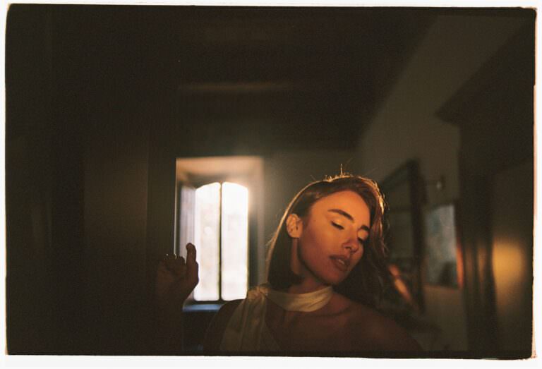 In soft sunlight filtering through a window, a woman in a white top with a shoulder wrap stands with eyes closed, evoking the timeless charm of an analog film in Italy. The dimly lit background, with blurred frames, creates a serene, contemplative atmosphere. Tuscan Wedding Photographer