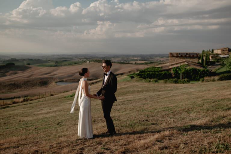 A couple in formal attire stands holding hands on a grassy hilltop during their elopement wedding at Capanna Suites Montalcino. The groom is in a black suit, and the bride wears a white gown. Rolling hills, a distant farmhouse, and a cloudy sky bathed in soft sunlight form the perfect backdrop. Tuscan Wedding Photographer