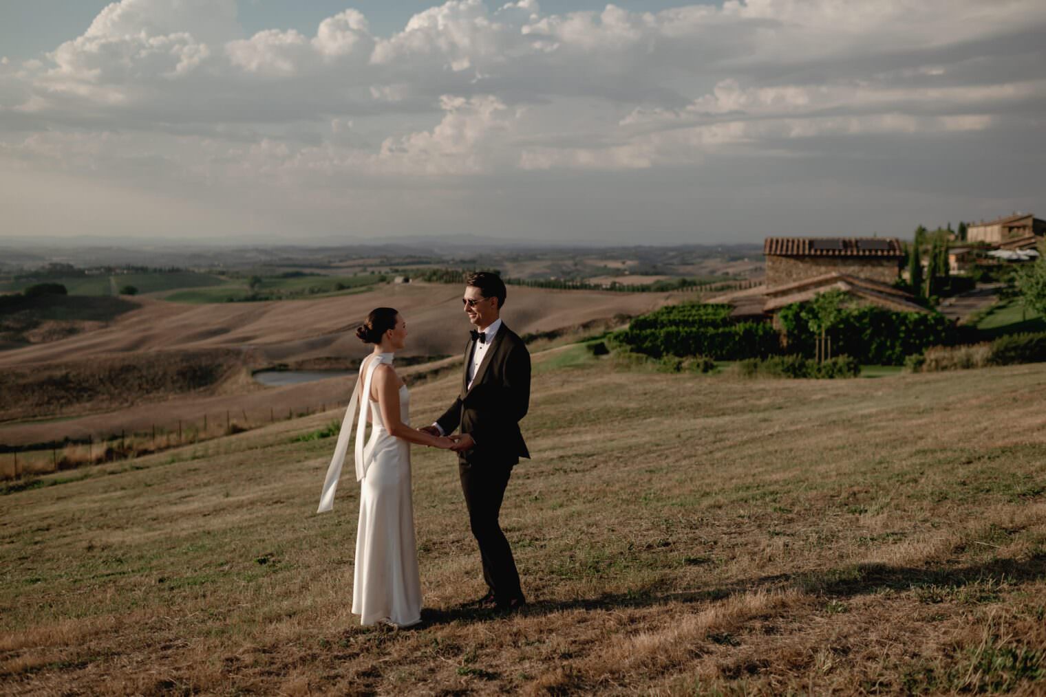 A couple in wedding attire holds hands on a grassy hillside in Tuscany, overlooking a vast landscape. The bride, in a white dress, and the groom, in a black suit, embrace the serene scene. Rolling hills and a rustic building complete this picturesque moment captured by their elopement photographer. Tuscan Wedding Photographer