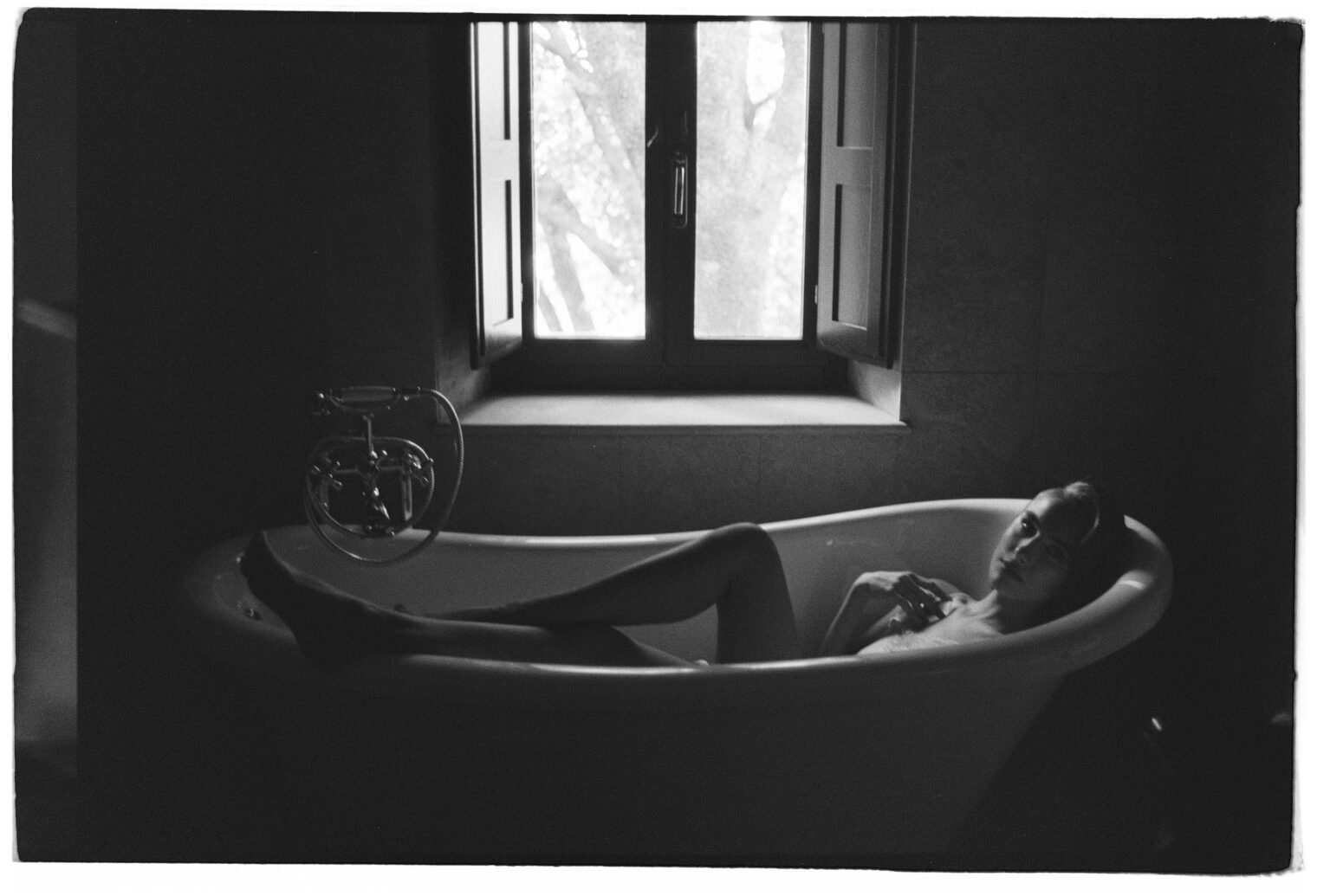 A black and white photo of a person reclining in a freestanding bathtub in a dimly lit bathroom. The window behind them shows a view of trees, with light softly illuminating the scene. The person appears relaxed, holding an object close to their chest. Tuscan Wedding Photographer