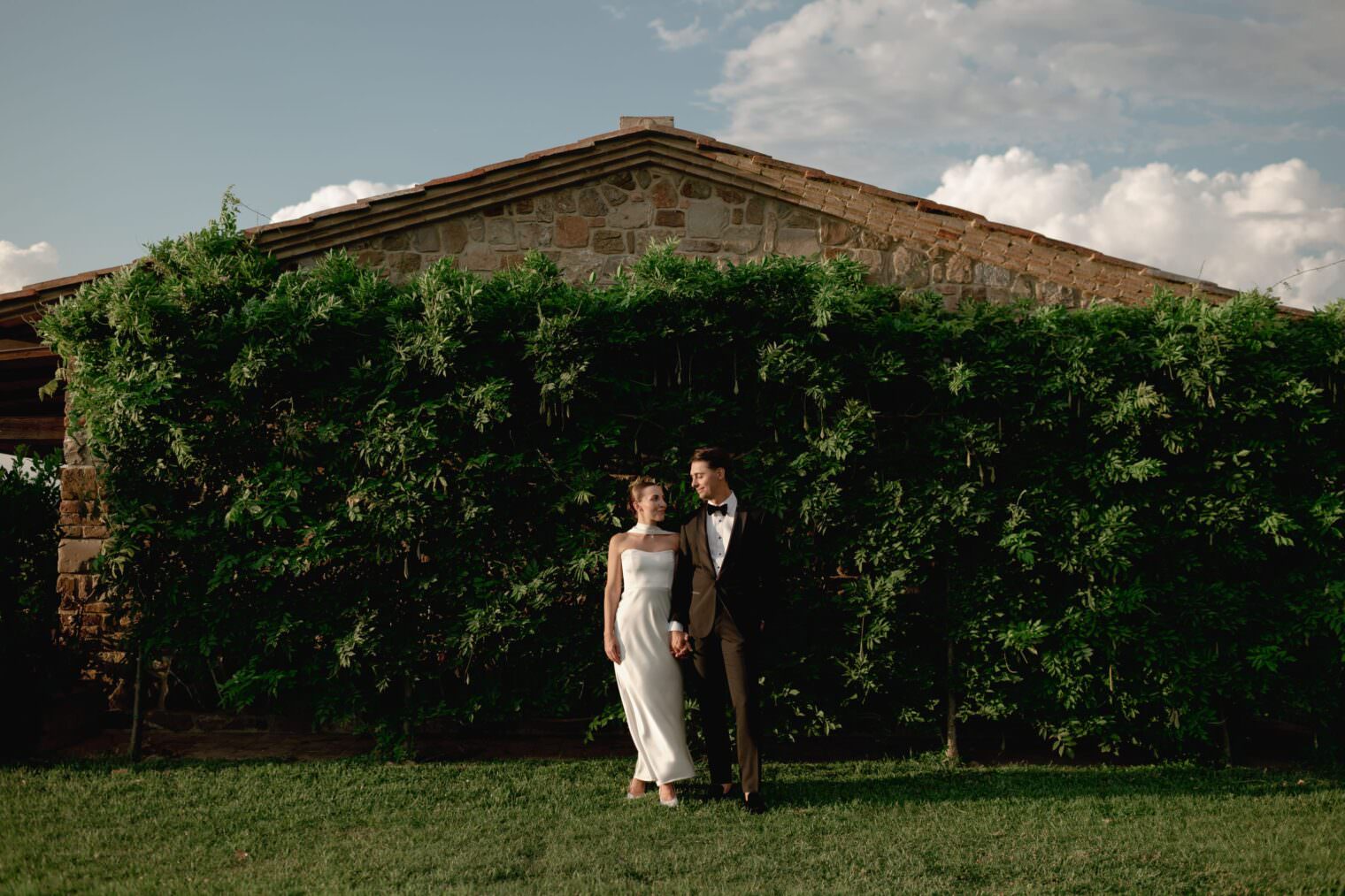 A bride in a white dress and a groom in a brown suit with a black bow tie stand hand in hand on a grassy lawn, captured by an expert wedding films photographer. Behind them, stone walls of an Italian villa peek through lush vines under a partly cloudy sky with soft, diffused light. Tuscan Wedding Photographer