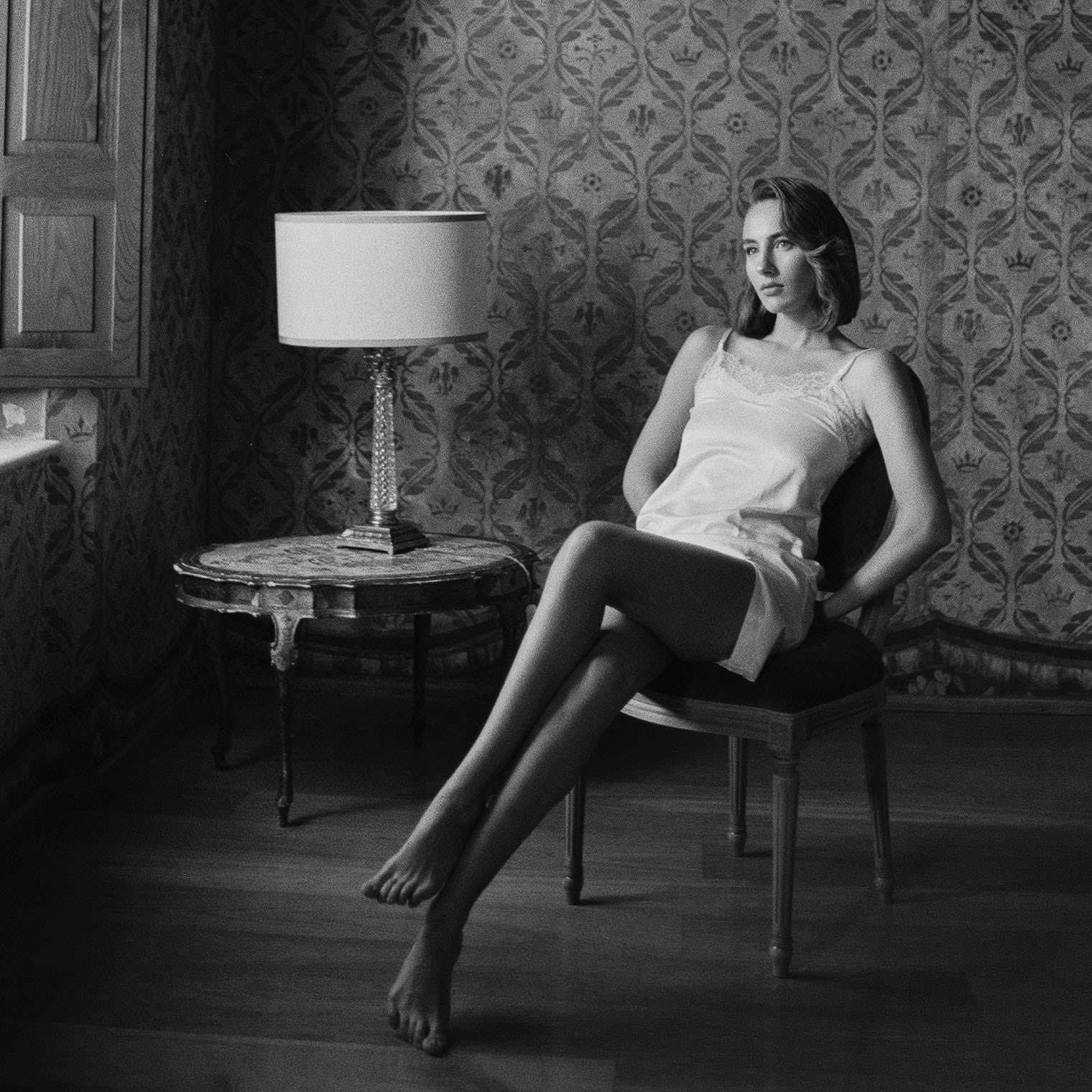 A woman in a white dress sits on a chair in an elegantly decorated room. Her legs are crossed, and shes looking thoughtfully to the side. The background features ornate wallpaper, and theres a lamp on a round table beside her. Natural light filters through a window on the left. Tuscan Wedding Photographer