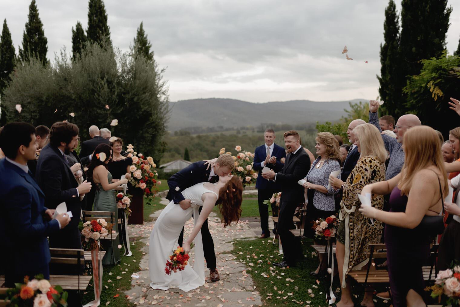 At their Palazzo Al Piano wedding, a couple kisses passionately amid a scenic outdoor aisle. The bride holds a bouquet of red and orange flowers. Guests, seated on wooden benches adorned with blooms, cheer and throw petals as tall trees and rolling hills form the picturesque background. Tuscan Wedding Photographer