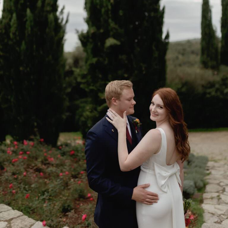A couple stands outdoors on a stone path, surrounded by greenery and flowers. The woman, with long red hair and a white dress featuring a large bow on the back, smiles at the camera. The man, in a dark suit, gently holds her waist, looking at her. Tall trees frame the background. Tuscan Wedding Photographer
