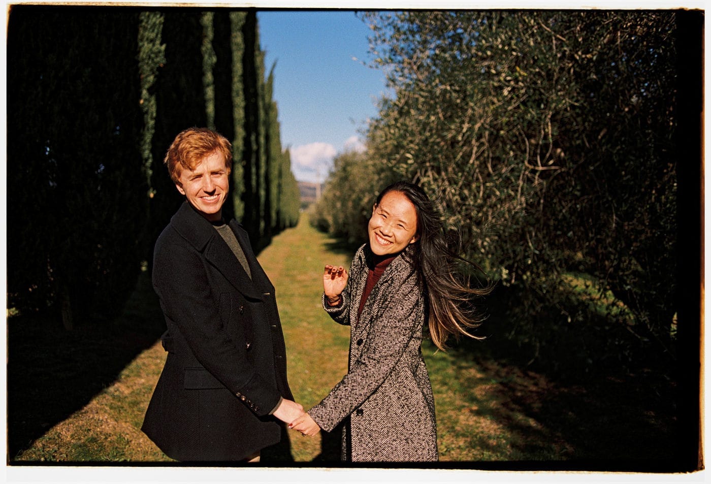 A man and woman joyfully hold hands outdoors on a sunny day. They are standing on a grassy path lined with tall trees. Both are wearing coats, and the woman’s hair is flowing in the breeze. The sky is clear and blue, creating a cheerful, serene atmosphere. Tuscan Wedding Photographer