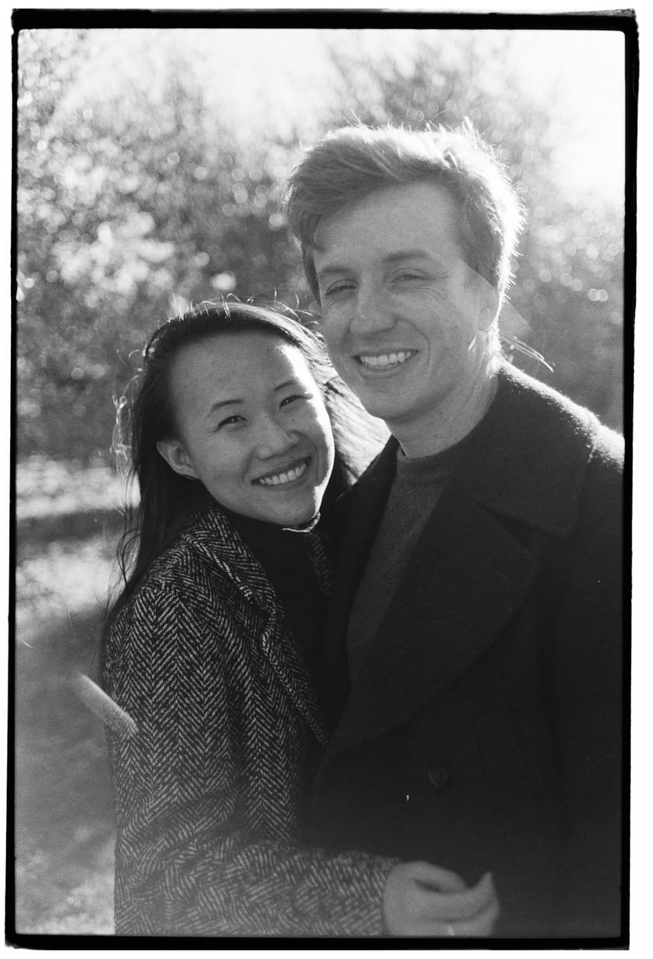 A black and white photo features a smiling couple outdoors. The woman is on the left, wearing a herringbone-patterned coat, while the man on the right is in a dark coat. Sunlight filters through trees in the background, highlighting the couple. Both are looking at the camera. Tuscan Wedding Photographer