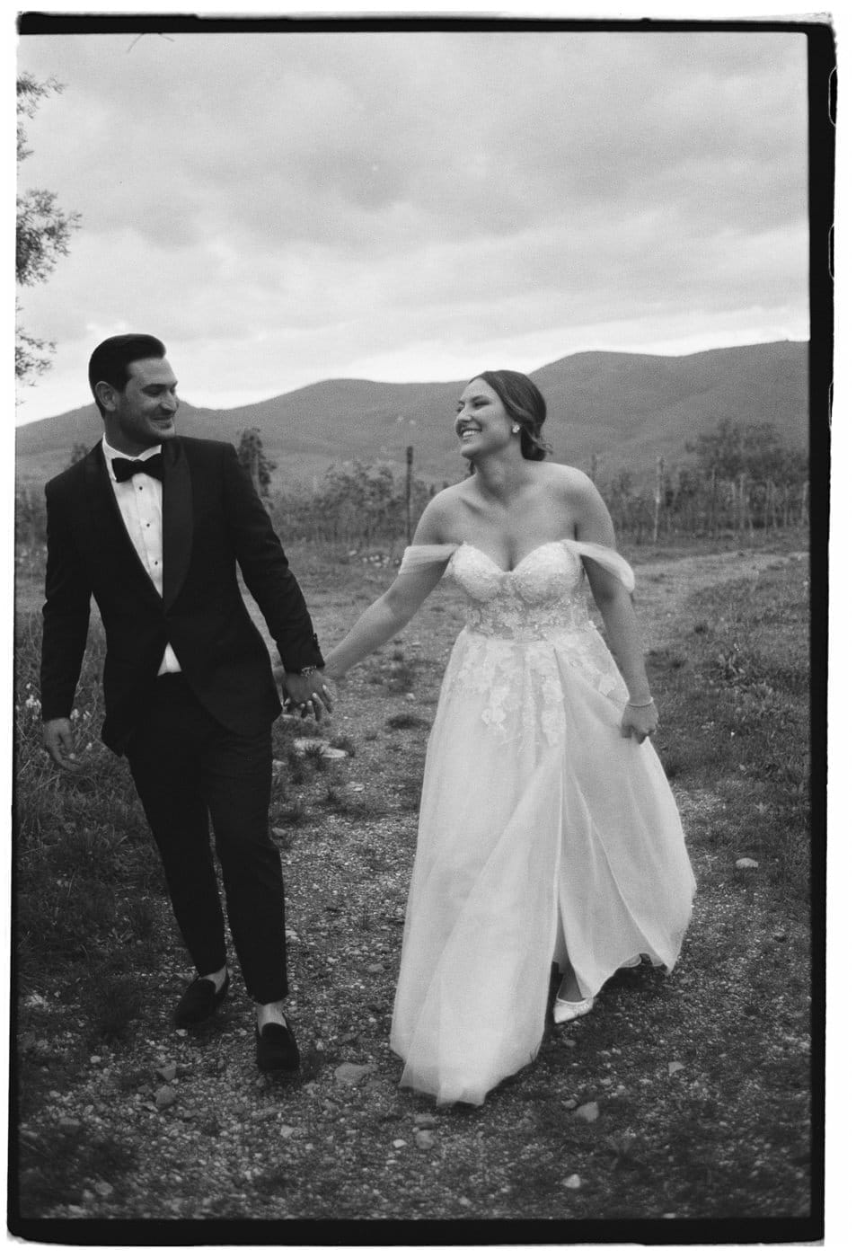 A bride and groom walk hand in hand in a rural outdoor setting. The bride wears a strapless, flowing wedding dress and the groom is in a tuxedo. They are smiling and looking at each other. The background features rolling hills and cloudy skies, creating a serene atmosphere. Tuscan Wedding Photographer