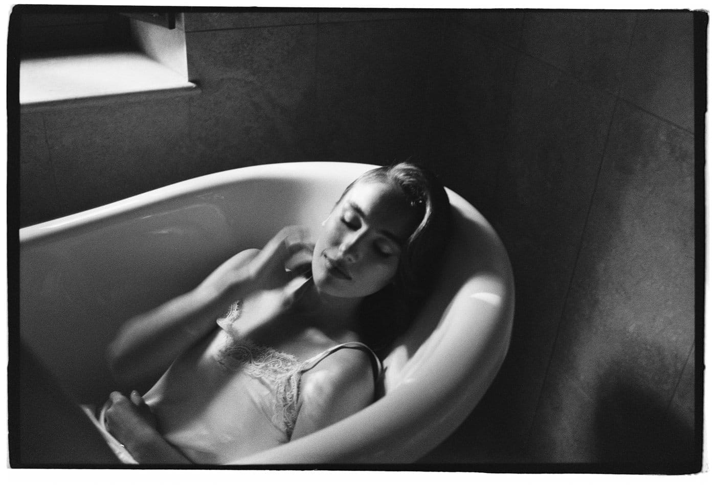 Black and white photo of a woman in a bathtub, wearing a camisole. Her eyes are closed, and her head rests on the tubs edge. Soft light enters through a nearby window, highlighting her serene expression and casting gentle shadows on the tiled walls. Tuscan Wedding Photographer