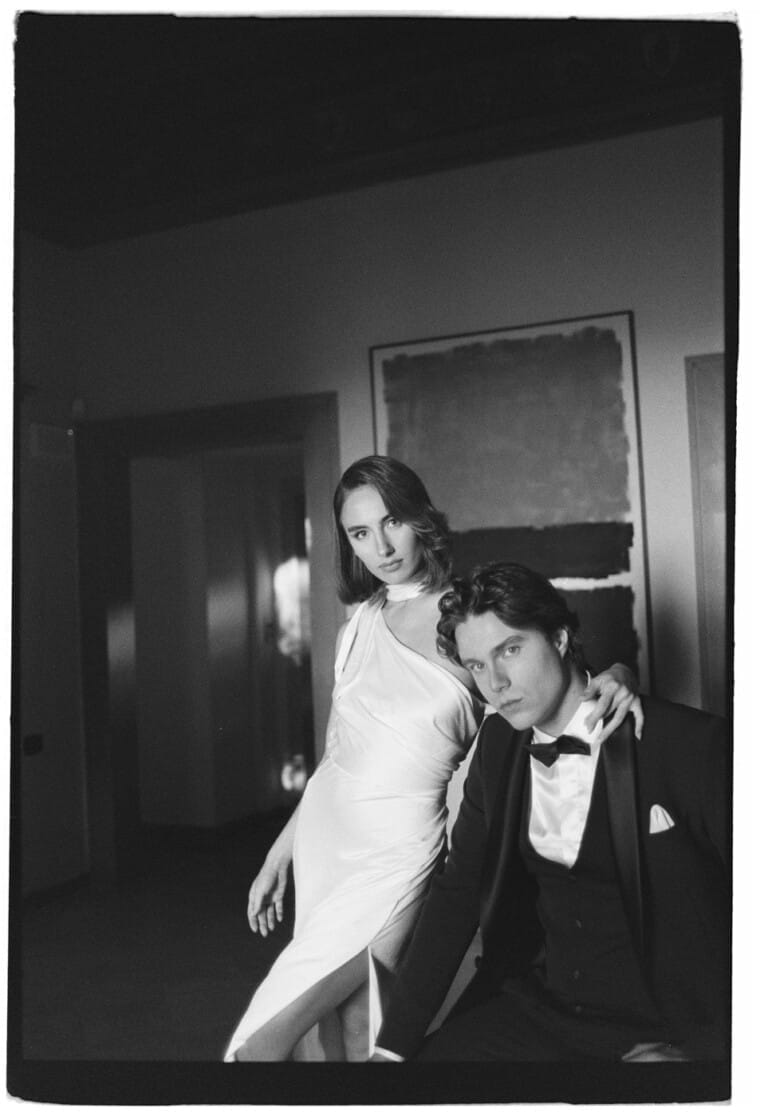 A black-and-white photo of a couple dressed elegantly. The woman stands in a flowing white gown with a high slit, draping her arm over the shoulder of a seated man in a tuxedo. They pose confidently, with a blurred artwork behind them and soft lighting enhancing the scenes sophistication. Tuscan Wedding Photographer