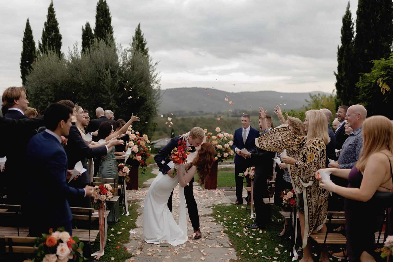 A bride and groom share a kiss at the end of an outdoor wedding aisle. Guests stand on either side, tossing flower petals in celebration. The couple is surrounded by greenery and floral arrangements. The background features a mountainous landscape under a cloudy sky. Tuscan Wedding Photographer