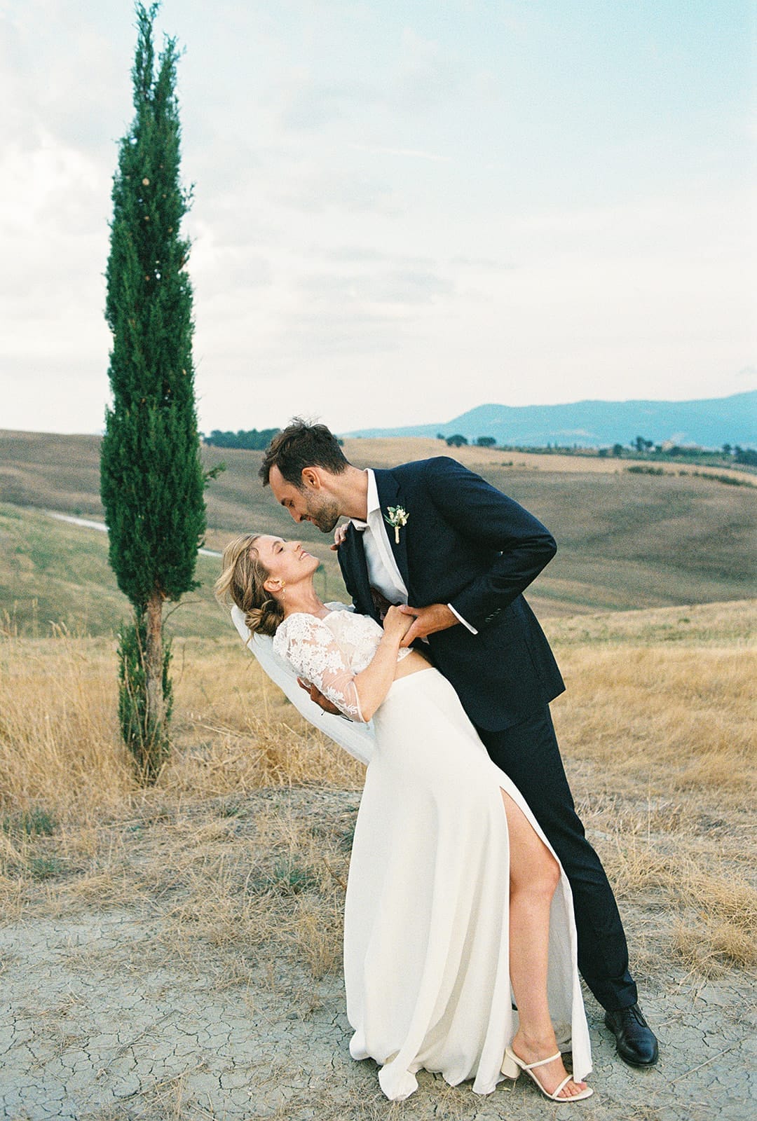 A couple is joyfully dancing outdoors. The man in a black suit leans over the woman in a white dress with lace sleeves. They embrace and gaze at each other, surrounded by dry grass and a tall, slender tree on a hillside. The sky is partly cloudy, and mountains are in the background. Tuscan Wedding Photographer