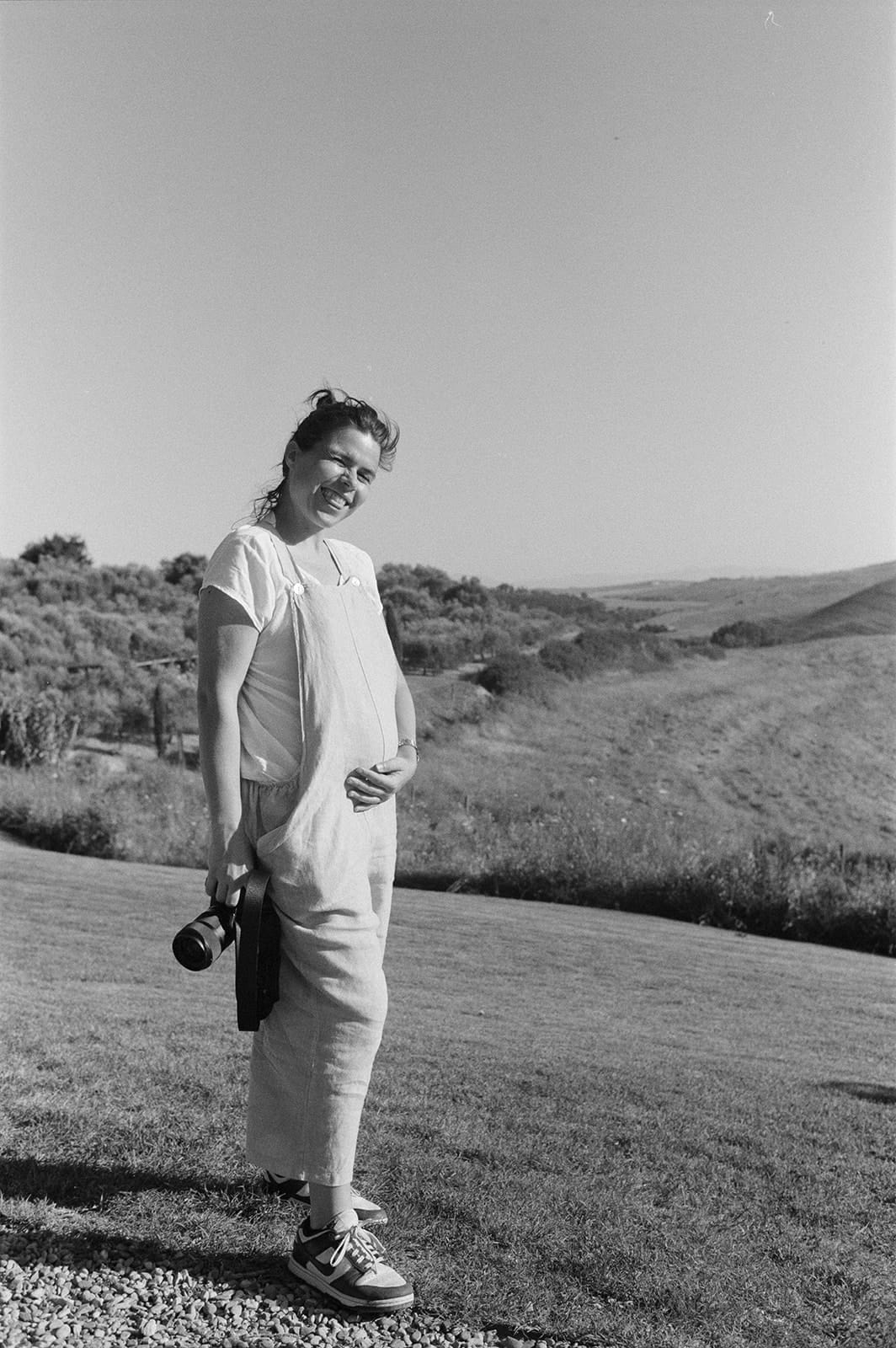 A woman stands outdoors on a grassy hill, holding a camera in one hand and resting the other on her abdomen. She wears light overalls, a white t-shirt, and sneakers. The background features rolling hills and clear skies, suggesting a serene, sunny day. The image is in black and white. Tuscan Wedding Photographer