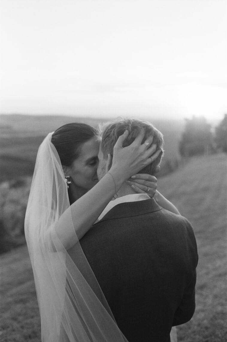 A black-and-white image of a couple embracing outdoors. The woman, wearing a veil and earrings, gently holds the mans head as they kiss. He is in a suit, facing away from the camera. The background features a blurred landscape with fields and a bright sky. Tuscan Wedding Photographer