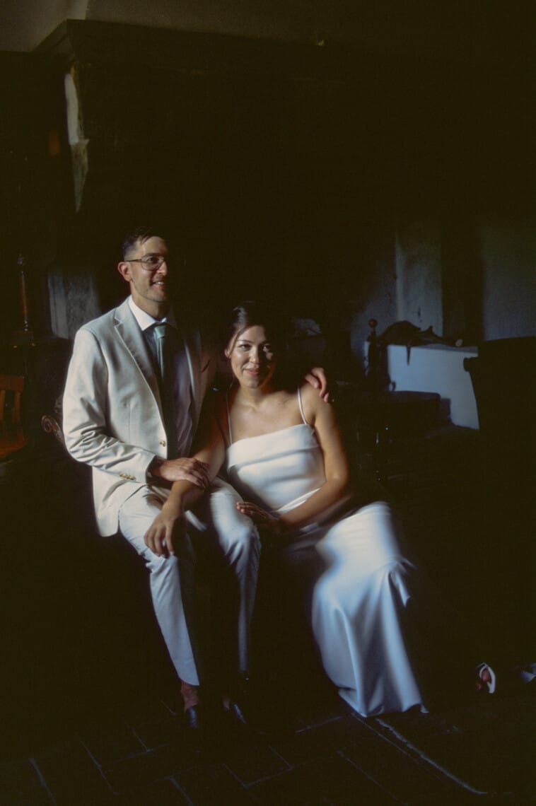 A couple poses indoors with dim lighting. The man, wearing a light suit and glasses, stands beside the seated woman, who is in a white dress. They both have a relaxed demeanor, with her holding his arm. The background features dark wooden furniture and a shadowed wall. Tuscan Wedding Photographer
