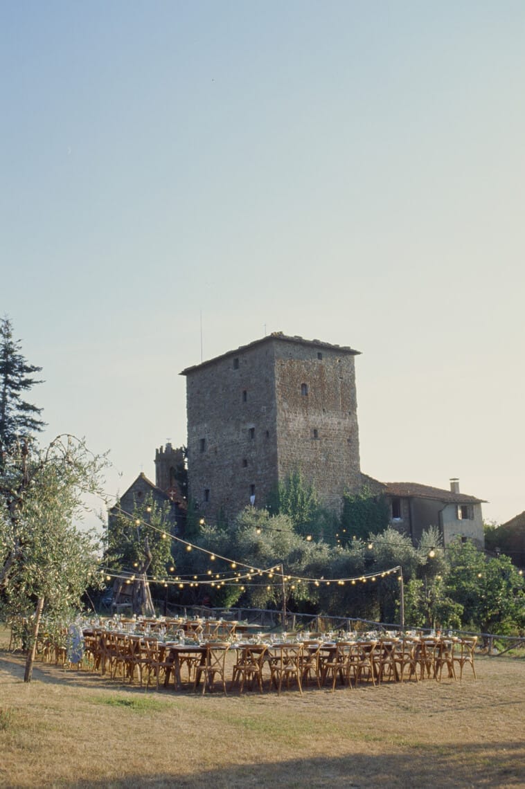 Outdoor wedding setup with wooden tables and chairs arranged in a garden. String lights are suspended above the tables. In the background, there is a tall, rustic stone building surrounded by trees and greenery. The sky is clear with soft, warm lighting. Tuscan Wedding Photographer