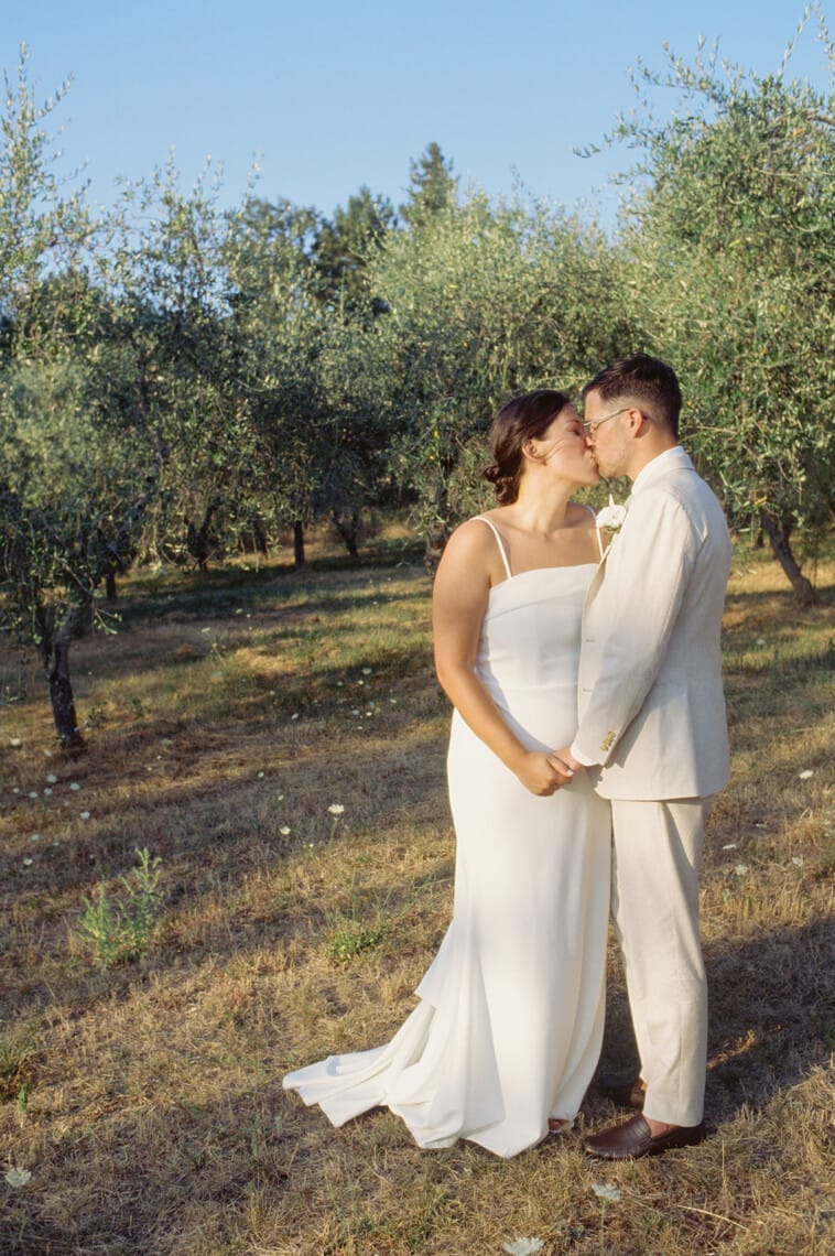 A couple stands in a sunlit grove, surrounded by olive trees. The woman wears a white dress, and the man is in a light beige suit. They are kissing, holding hands, and barefoot on the grassy ground. The scene is serene and romantic, with a clear blue sky above. Tuscan Wedding Photographer