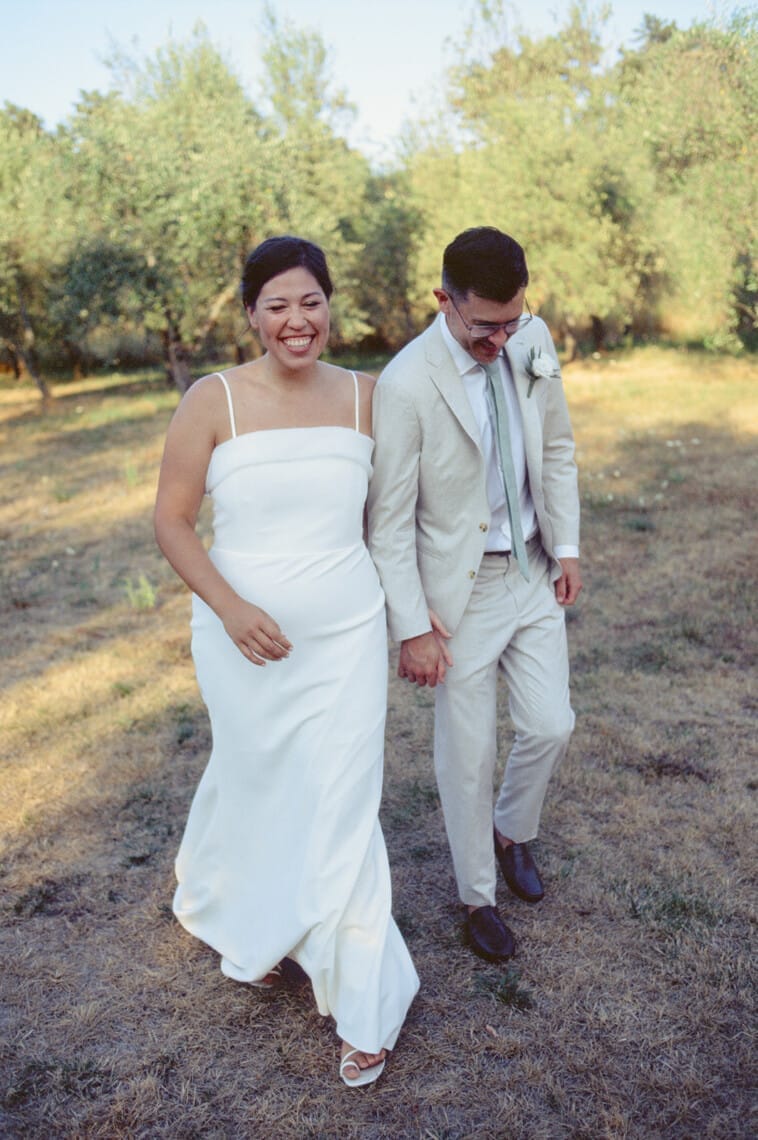 A joyful couple walks hand in hand outdoors on grass. The woman wears a white dress, and the man wears a light-colored suit with a tie and boutonniere. Sunlight filters through the surrounding trees, creating a warm, serene atmosphere. Both are smiling and looking down. Tuscan Wedding Photographer