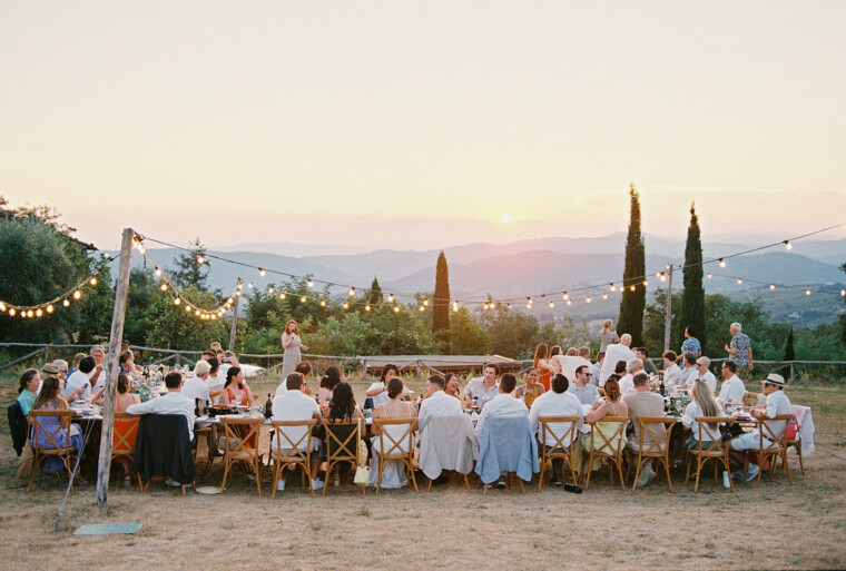 A large group of people seated around long tables at an outdoor event during sunset. The tables are decorated, and strings of lights are overhead. Attendees are dressed in summer attire. The background features rolling hills and cypress trees under a colorful sky. Tuscan Wedding Photographer