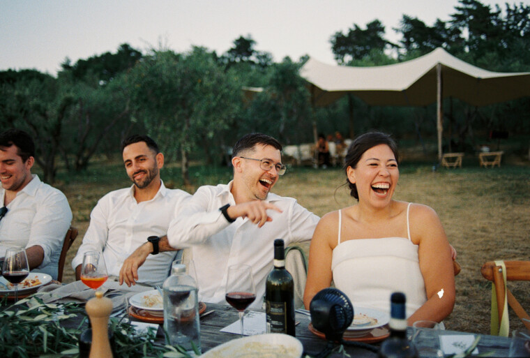 A group of people sitting at an outdoor table, laughing and enjoying a meal. They are dressed in semi-formal attire. The table is set with plates, wine glasses, and a wine bottle. In the background, trees and a beige canopy are visible under a clear sky. Tuscan Wedding Photographer