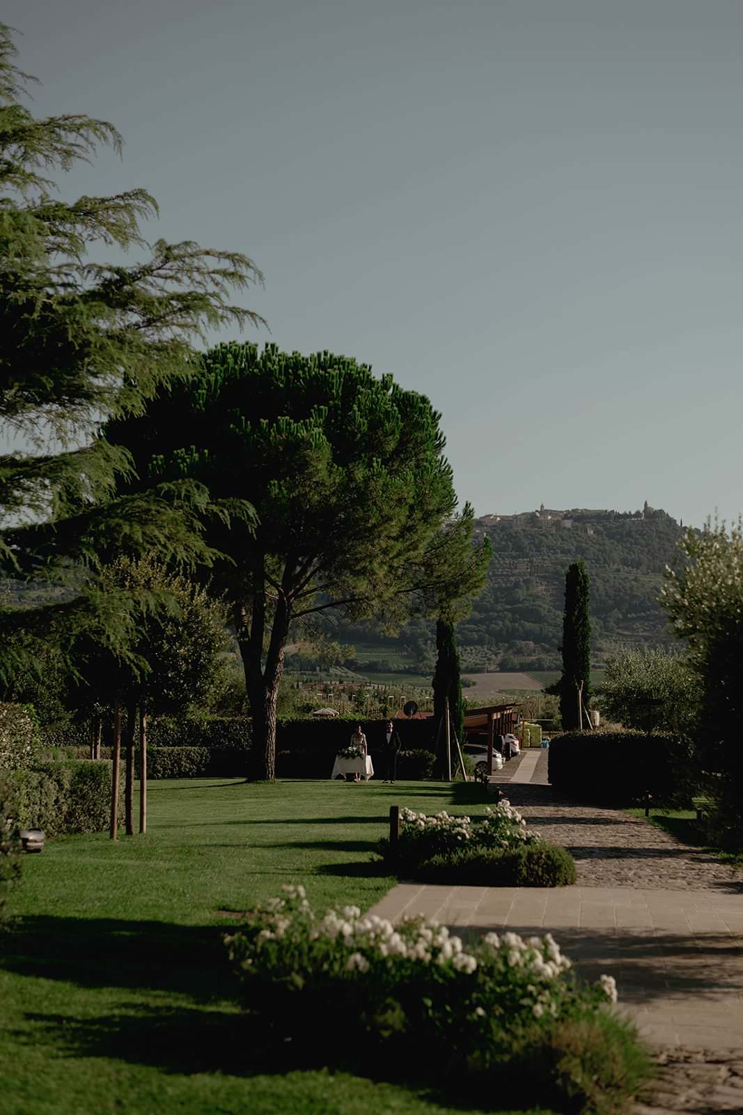 A serene outdoor scene features a tree-lined path leading to tables set for dining on a well-manicured lawn. Tall trees provide shade, while rolling hills and a distant town are visible under a clear blue sky. White flowers border the path, adding a touch of elegance. Tuscan Wedding Photographer