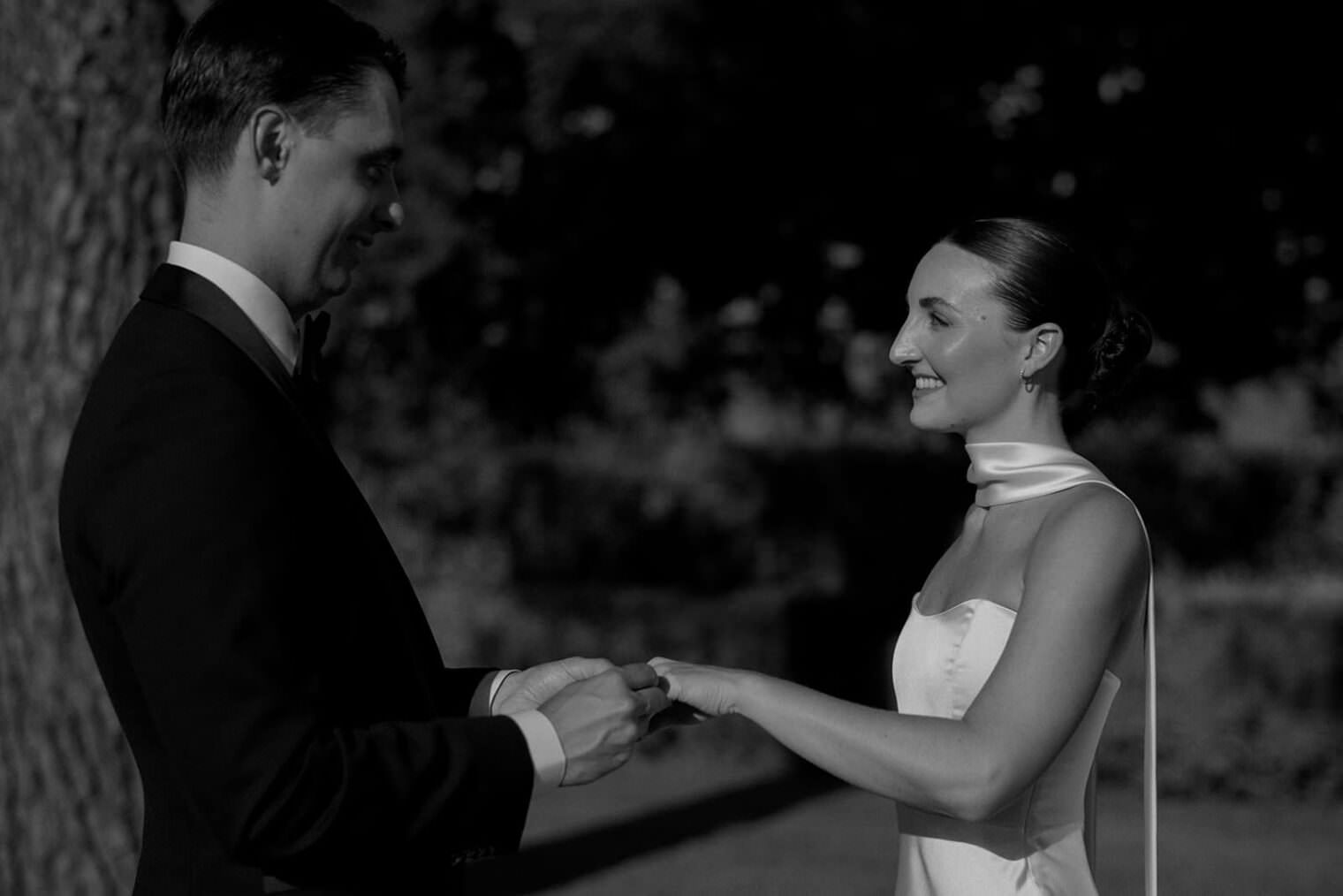 A couple stands facing each other outdoors in a black and white photo. The man, in a dark suit, gently holds the womans hand. The woman wears a white dress with a scarf and has her hair tied back. They appear poised, with trees and sunlight filtering in the background. Tuscan Wedding Photographer