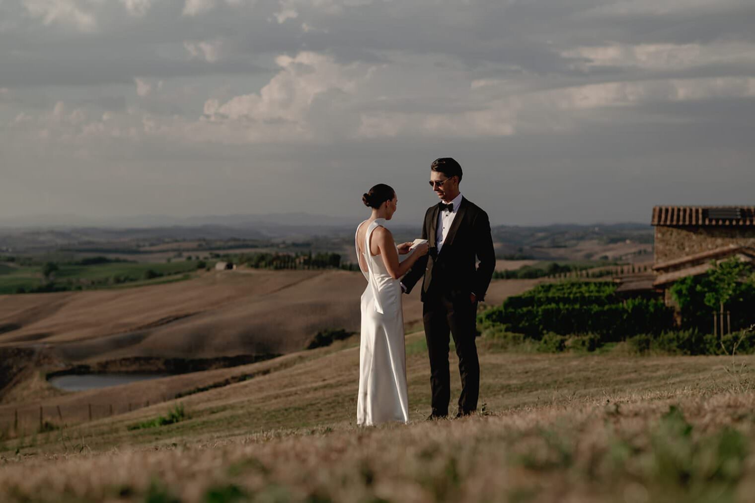 A couple stands on a grassy hill in elegant attire, with the woman in a white dress and the man in a black suit. They are exchanging rings or vows. The background features rolling hills under a cloudy sky, and a rustic building on the right enhances the scenic landscape. Tuscan Wedding Photographer