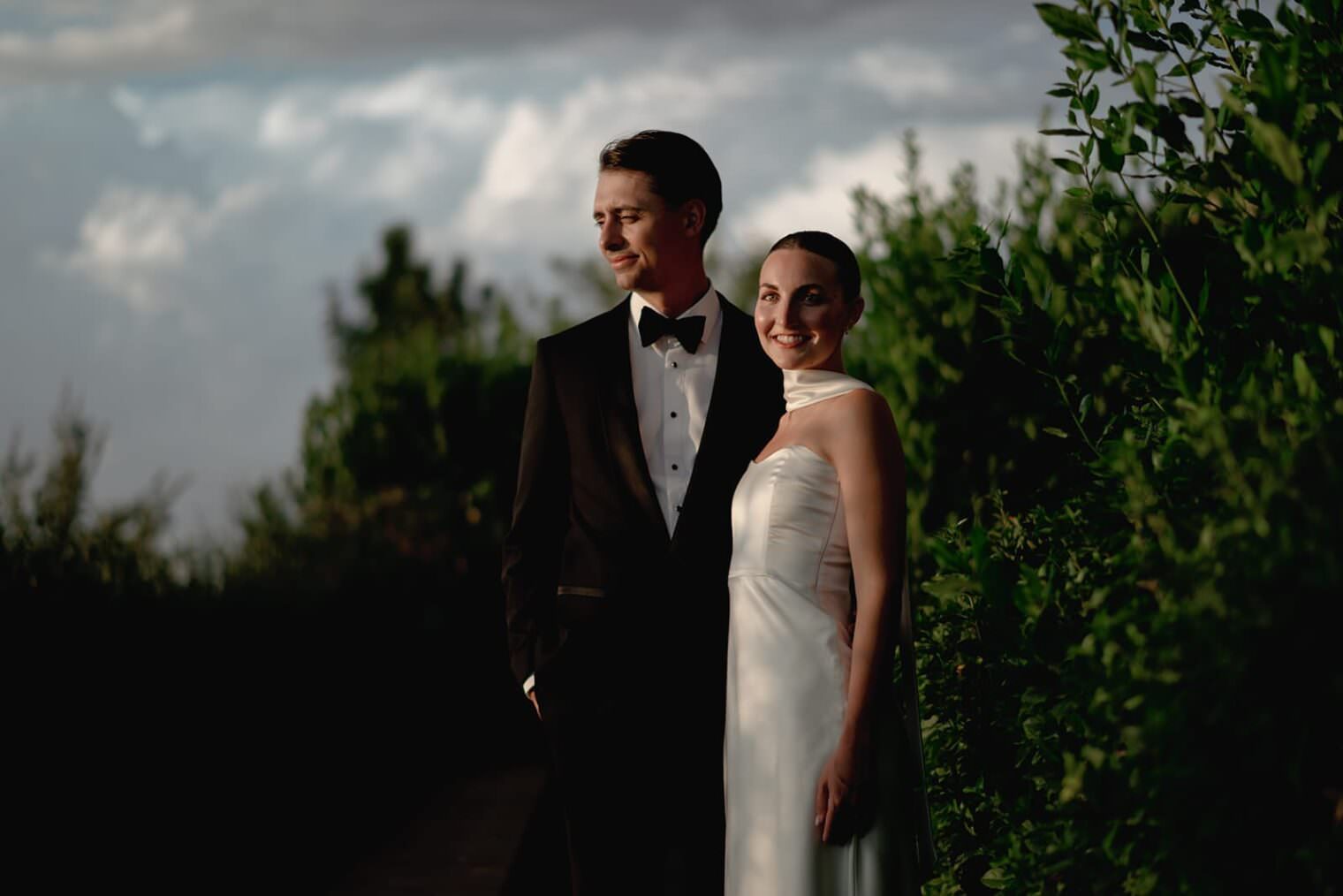 A couple stands outdoors, dressed formally. The man wears a black tuxedo and bow tie, looking to the left. The woman in a white sleeveless gown smiles at the camera. They are surrounded by lush greenery under a partly cloudy sky, with soft lighting creating a serene atmosphere. Tuscan Wedding Photographer