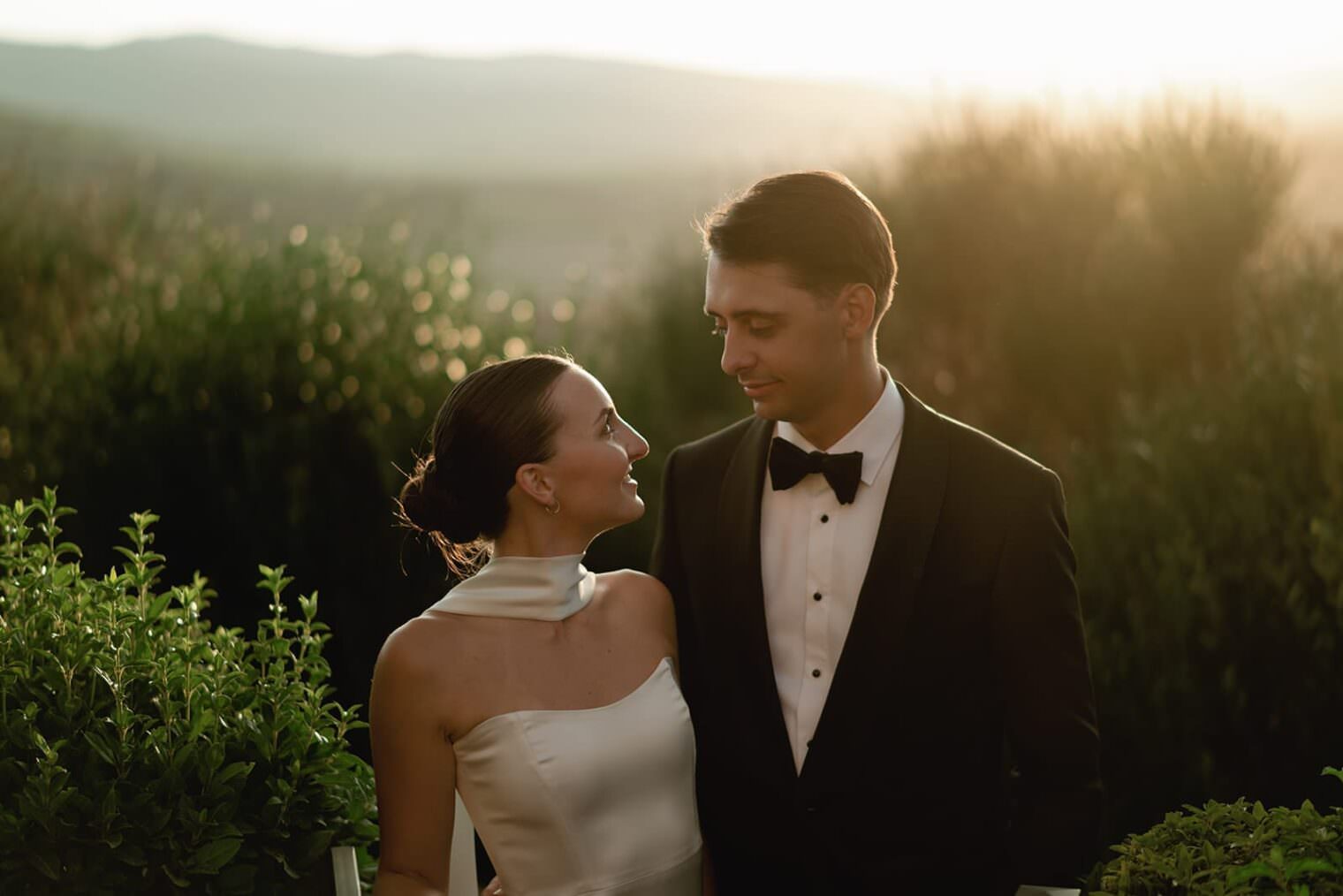 A couple stands together at sunset. The woman, in a sleeveless white gown with a high neckline, looks fondly at the man, who wears a black tuxedo and bow tie. They stand in a lush garden with blurred greenery and hills in the background, bathed in warm, golden light. Tuscan Wedding Photographer