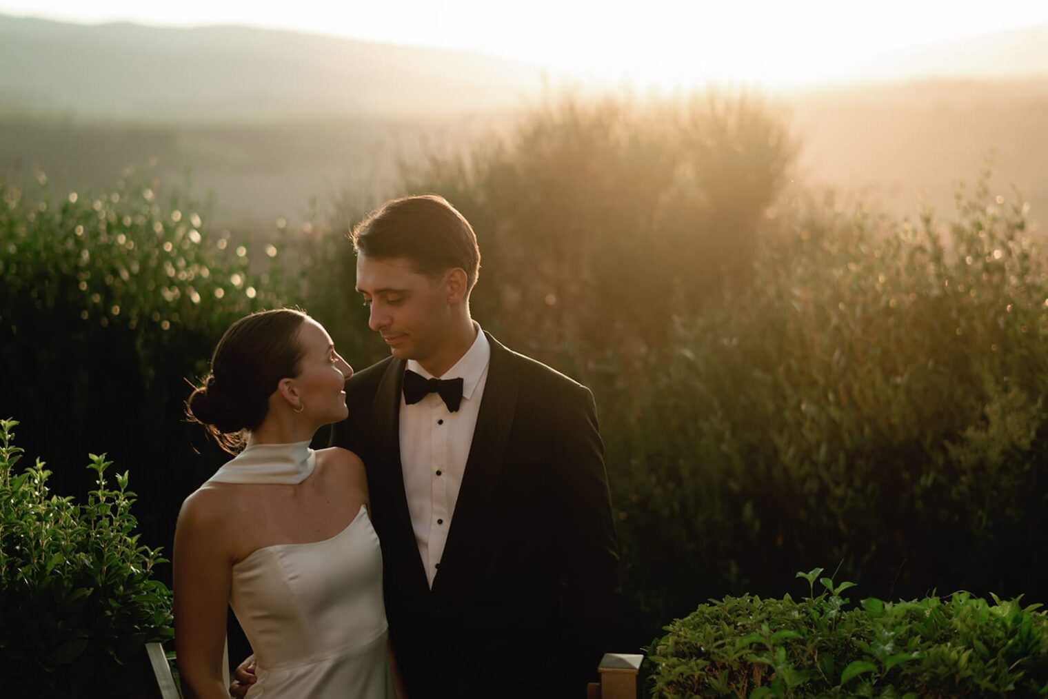 A couple stands outdoors at sunset. The woman wears a white halter-neck dress, and the man wears a black tuxedo with a bow tie. They gaze lovingly at each other, surrounded by greenery and softly lit by the sun setting over distant hills. Tuscan Wedding Photographer