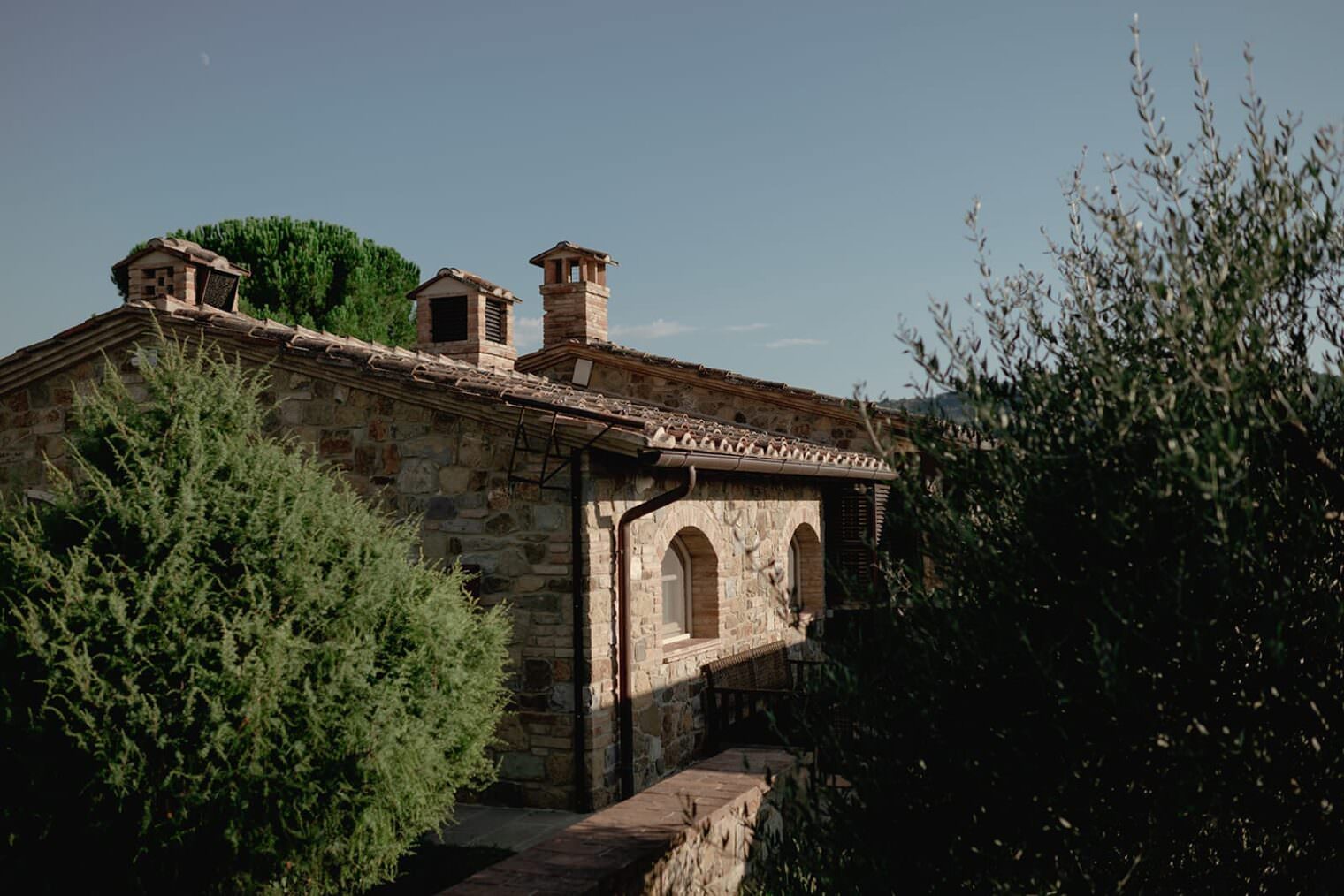 Nestled in the heart of Tuscany, this stone house with a rustic clay-tiled roof and small chimneys is a picturesque setting for elopement photos. Surrounded by lush green bushes and olive trees, the charming arches and wooden shutters glow under the clear blue sky on a sunny day. Tuscan Wedding Photographer
