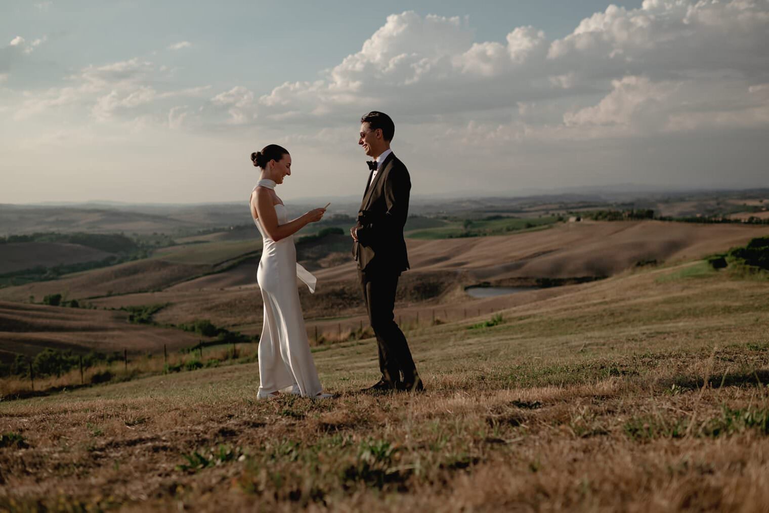 A couple stands joyfully in an open field with rolling hills and a cloudy sky. The woman, in a white dress, faces a man in a black suit and bow tie. As their wedding photographer captures this elopement, they hold hands on dry grass, creating an intimate scene under soft, warm lighting. Tuscan Wedding Photographer