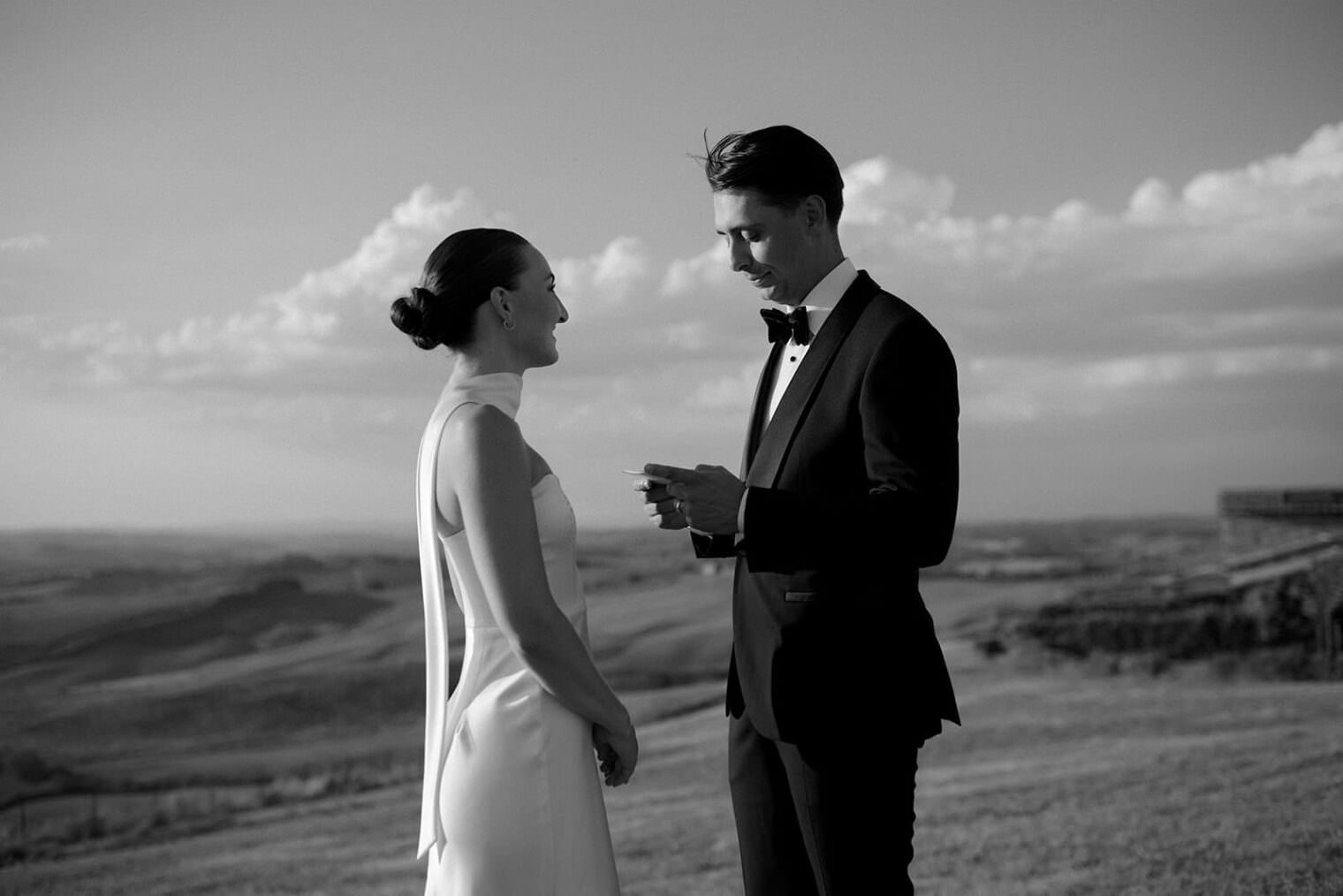 A bride and groom stand outdoors in an intimate elopement ceremony, silhouetted against Tuscanys rolling hills and a cloudy sky. The groom, in a tuxedo, reads from a notecard while the bride, in a sleeveless gown, gazes at him. Captured by a wedding photographer in timeless black and white. Tuscan Wedding Photographer
