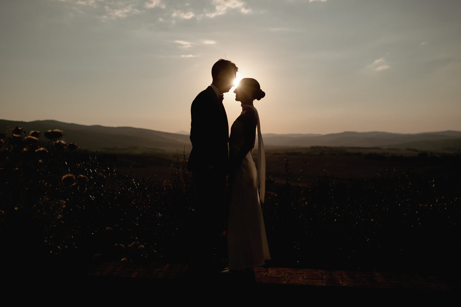 Silhouetted against a setting sun, a couple stands closely together on a brick overlook with rolling hills in the background. The sky is a gradient of warm colors, and wildflowers are visible on the left. The couple appears to be sharing an intimate moment amidst the tranquil landscape. Tuscan Wedding Photographer