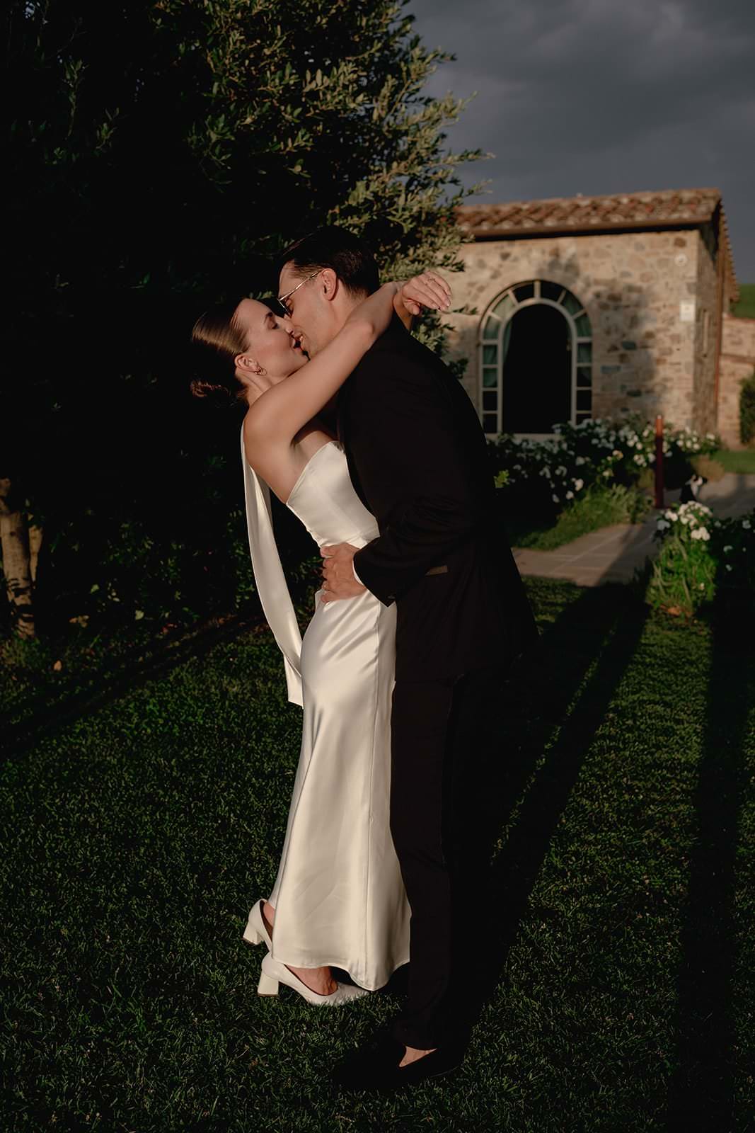 A couple shares a kiss in a garden during golden hour. The woman, in a sleek white satin dress, embraces the man wearing a black suit. Sunlight casts long shadows on the grass. A stone building with arched windows is visible in the background. Tuscan Wedding Photographer