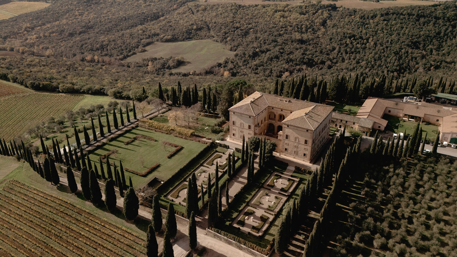 Aerial view of an Italian villa surrounded by lush greenery and vineyards. The villa features classic architecture with a rectangular layout, flanked by formal gardens and rows of tall cypress trees. Hills and dense forest create a picturesque backdrop under a clear sky. Tuscan Wedding Photographer