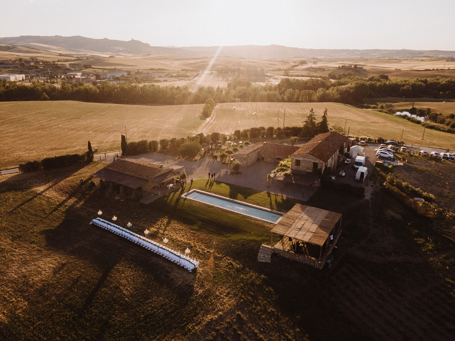 Aerial view of a sunlit countryside estate with rustic buildings, a narrow pool, and a long outdoor table setup. The surrounding fields are expansive, with lush greenery. In the distance, gentle hills and scattered trees are visible under a soft evening sky. Tuscan Wedding Photographer